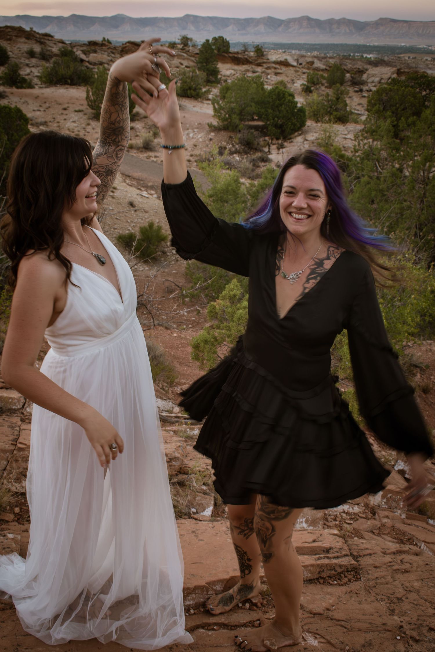 Two brides dancing outdoors in desert landscape, one in white gown and one in black dress with purple hair.