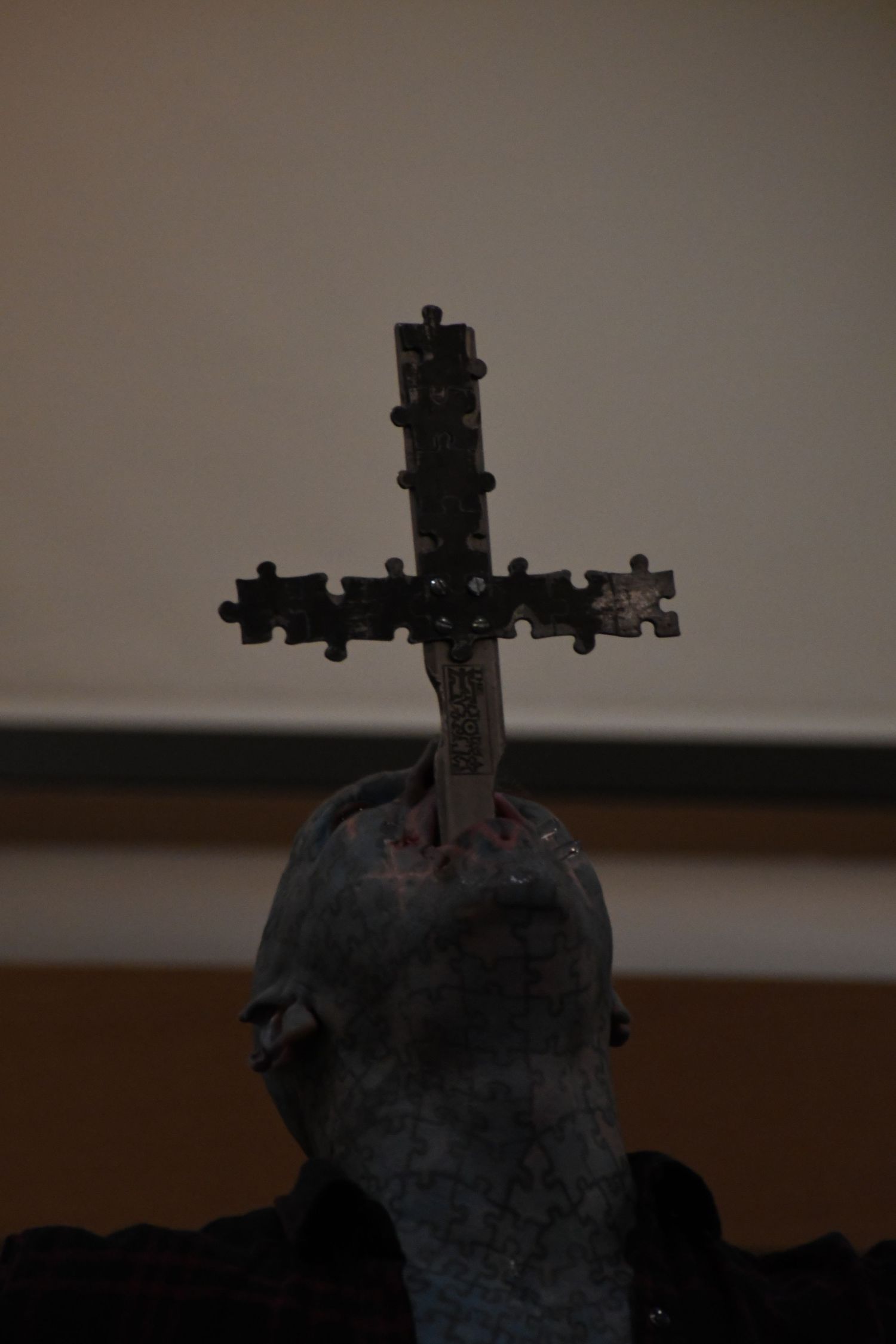 A dark metal cross adorned with puzzle pieces mounted on a sculptural base in a dimly lit room.
