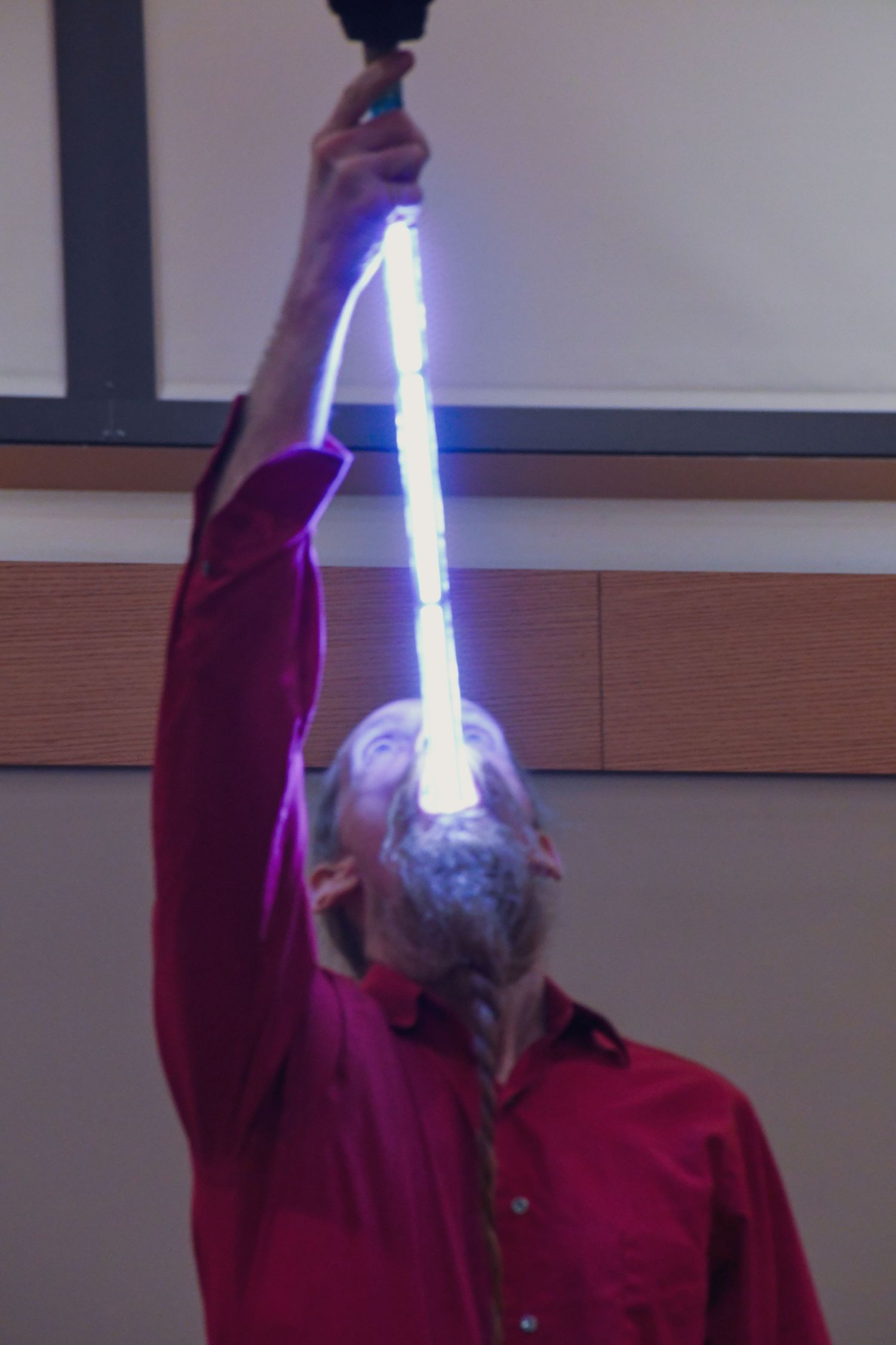 Man in red shirt holding glowing fluorescent light tube above his head in a darkened room.