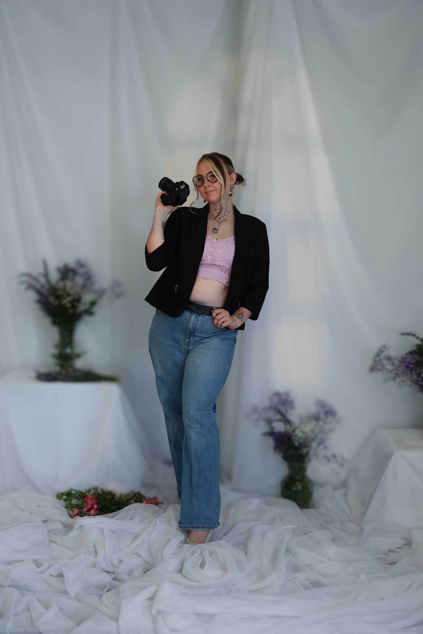 Blonde woman in black blazer and jeans holds camera to face before white draped floral backdrop.
