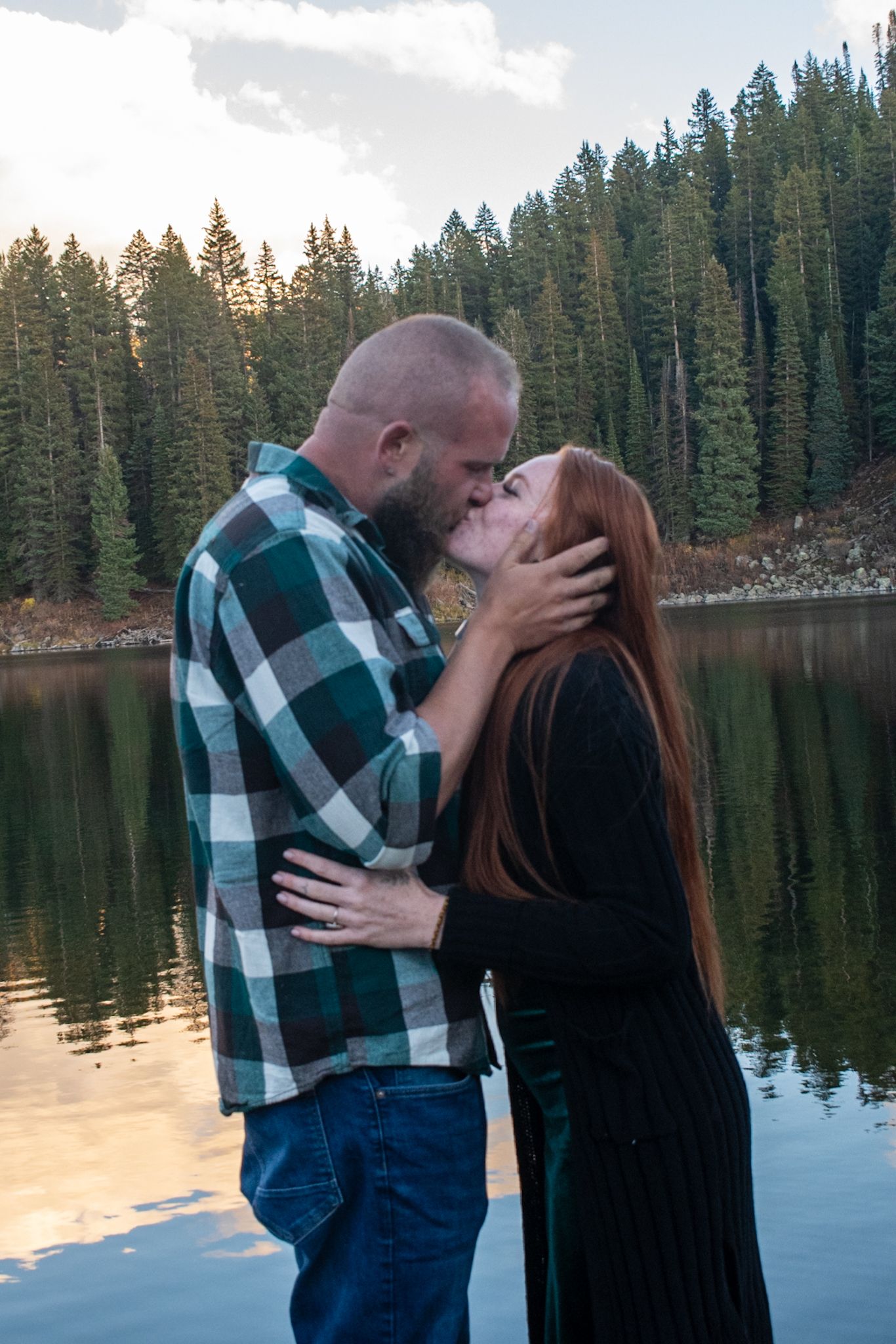 Couple sharing a romantic kiss by a reflective mountain lake surrounded by evergreen forest at sunset.