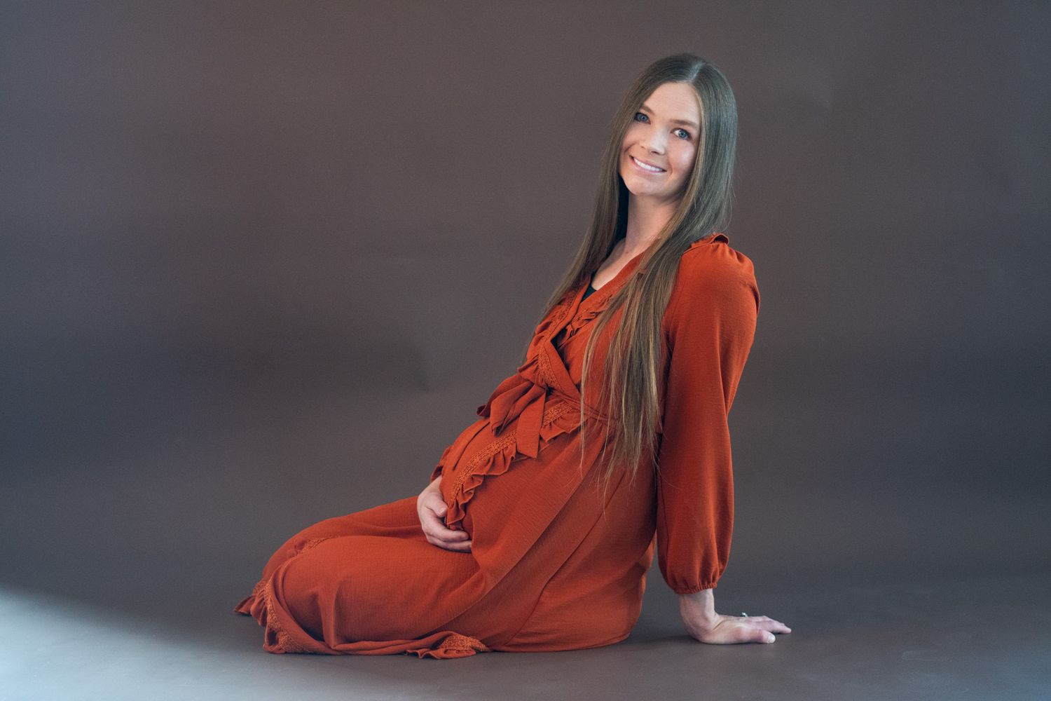 Smiling pregnant woman in red dress sitting on floor, long hair, studio photo shoot with gray background.