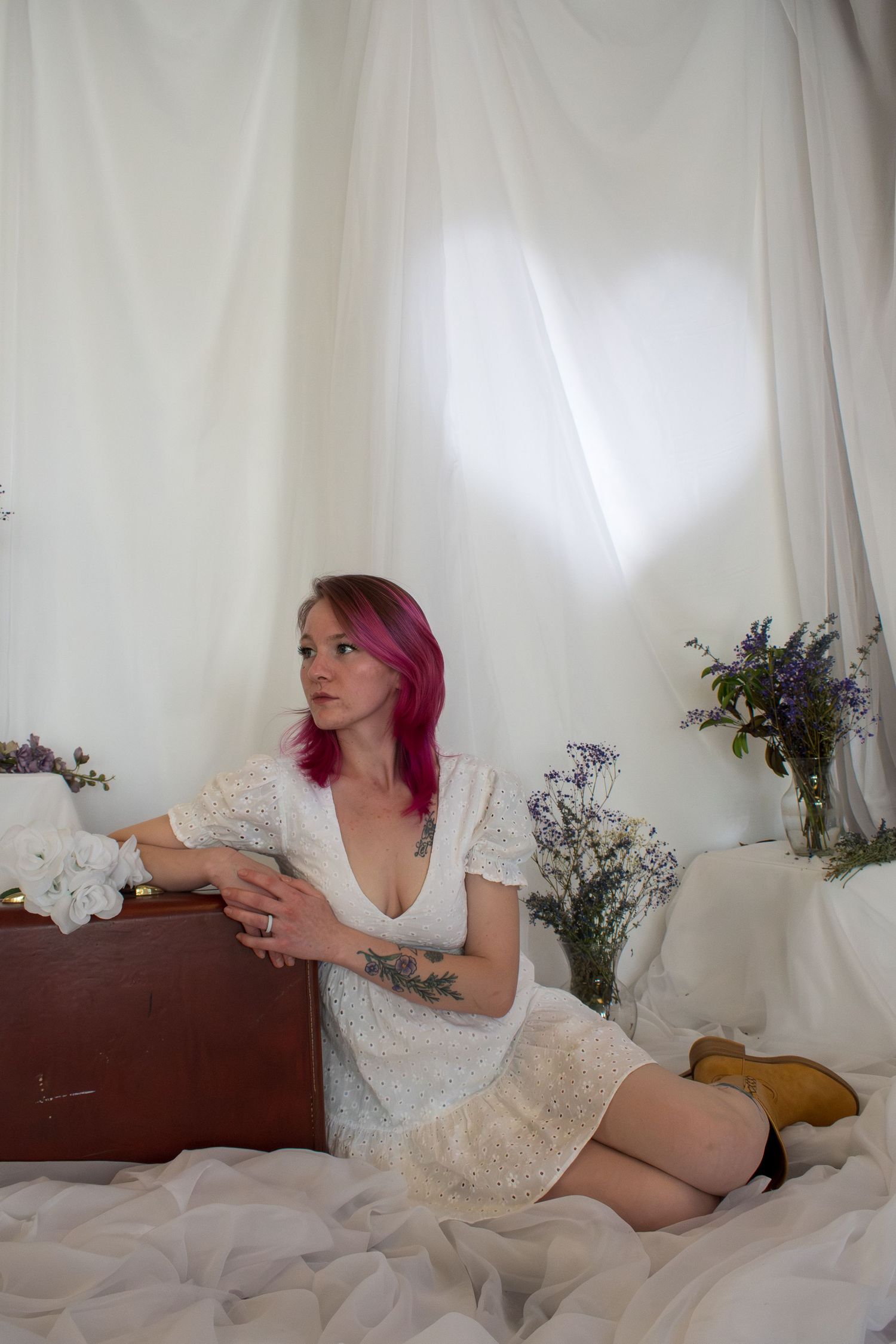 Young woman with pink hair in white lace dress sits beside vintage suitcase surrounded by wildflowers against white draping.