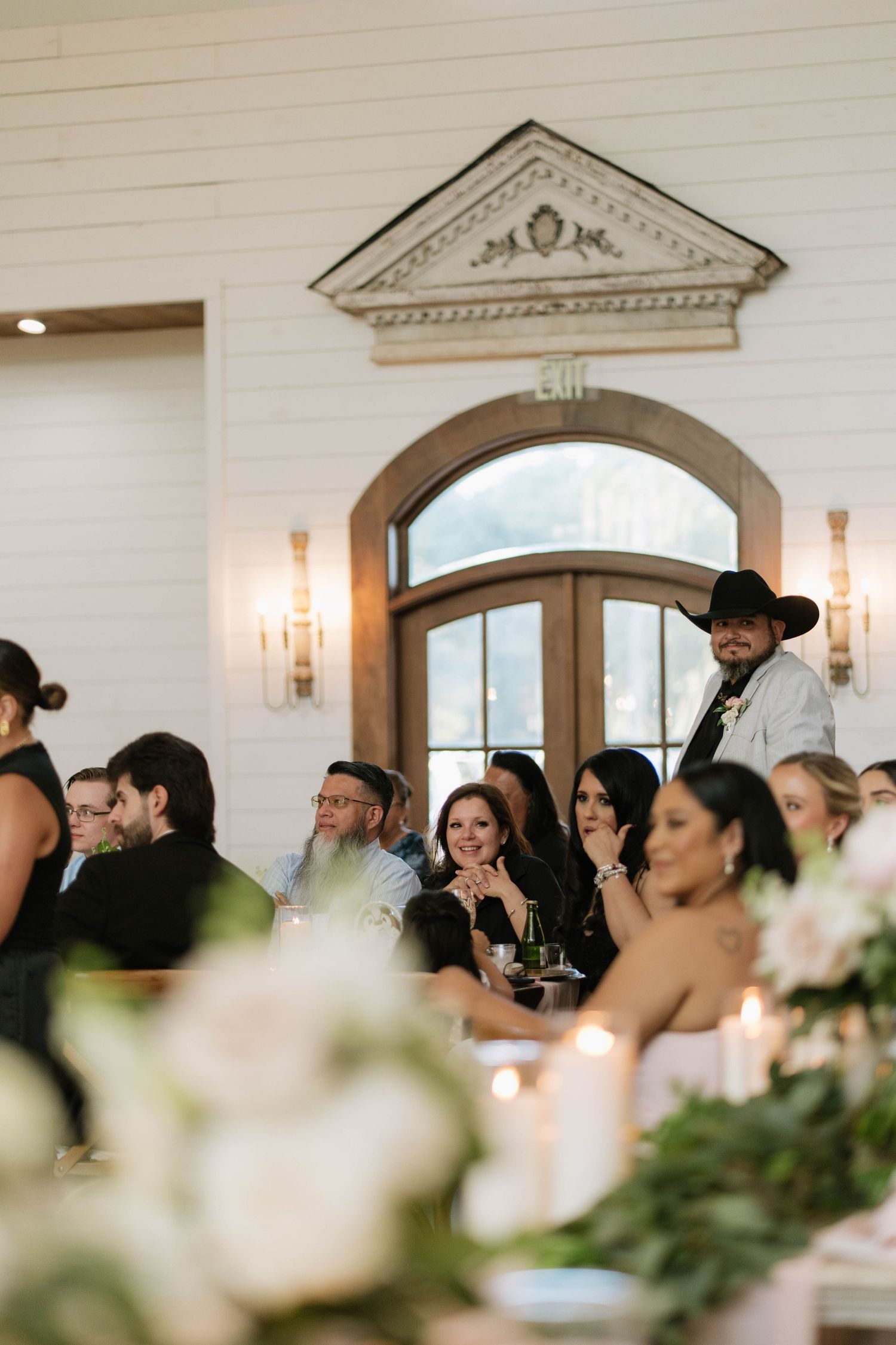 Wedding reception guests seated at elegant tables with white linens and greenery decor in a bright indoor venue.