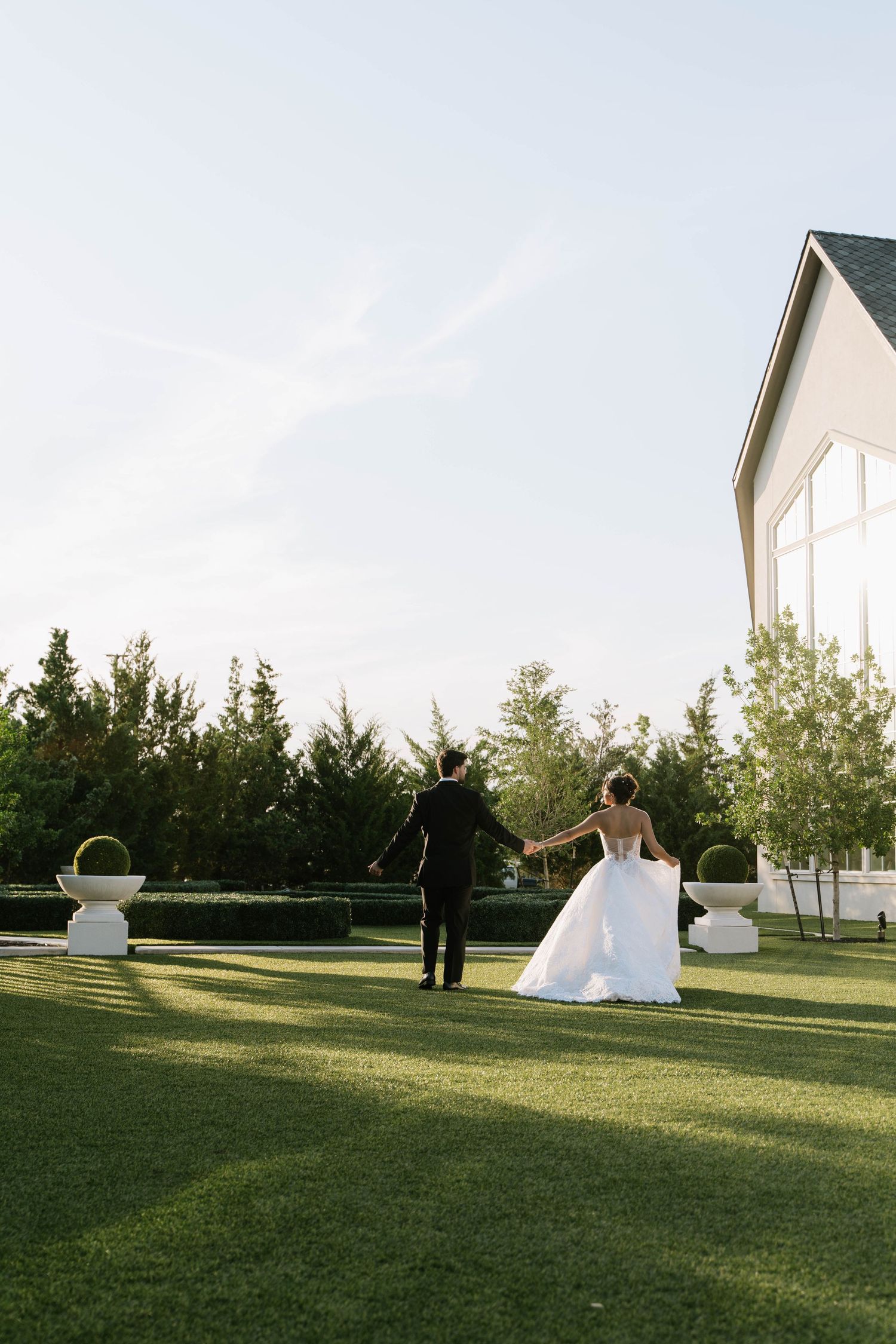 Bride and groom walk hand-in-hand across lush green lawn outside elegant white wedding venue.