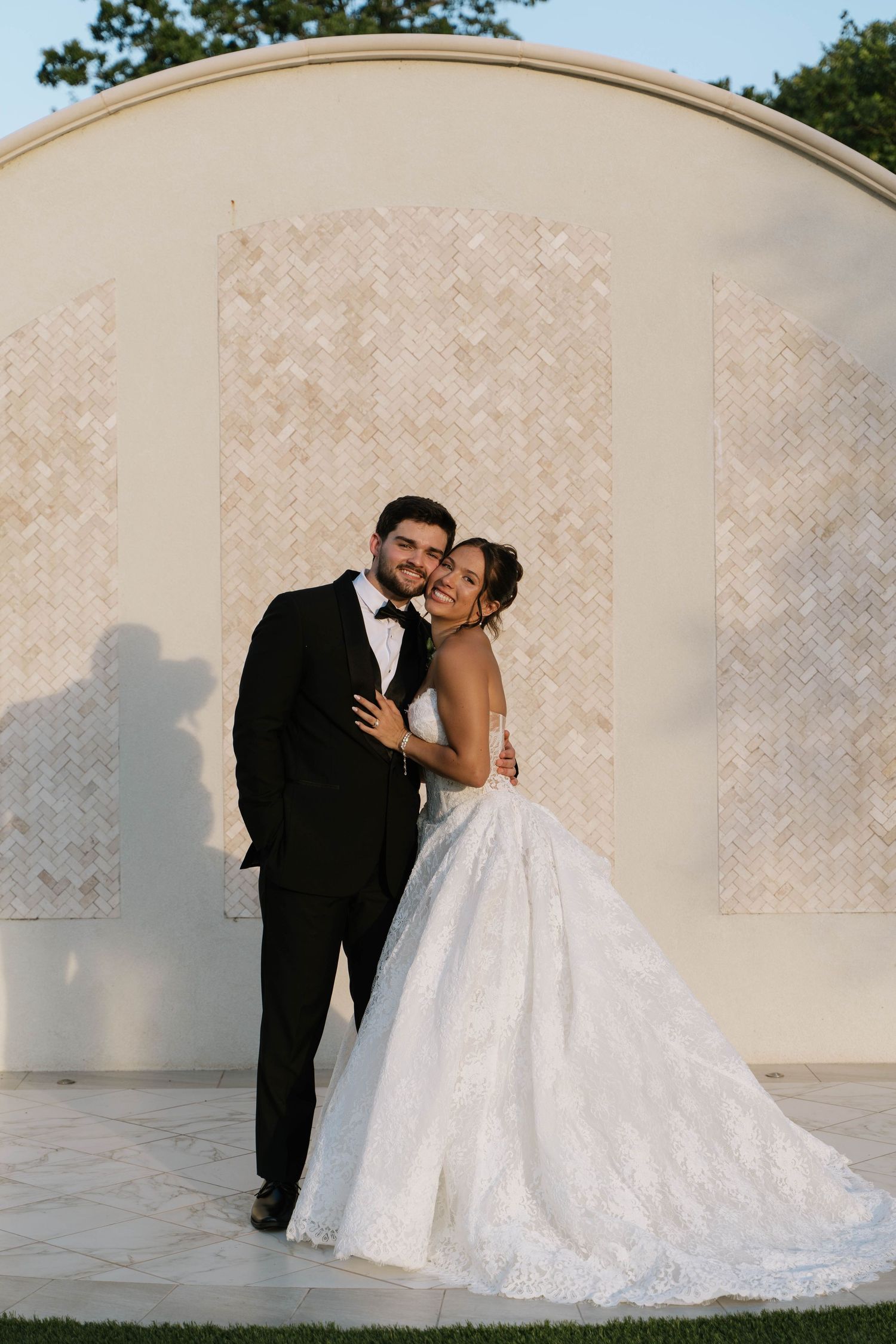 Bride in lace ball gown and groom in black tuxedo pose together in front of elegant herringbone stone wall.