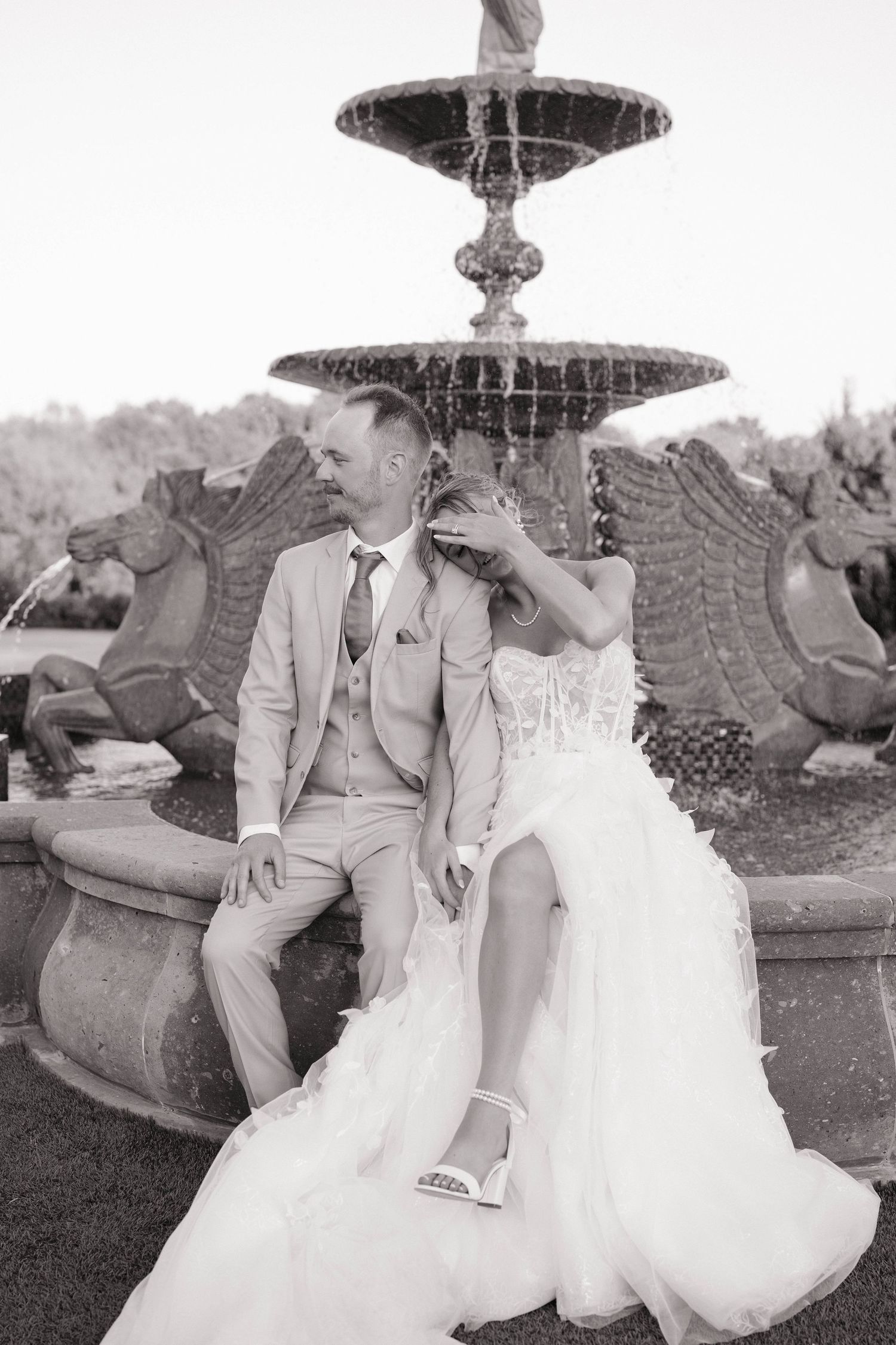 Black and white photo of a couple in wedding attire sitting by an ornate fountain.