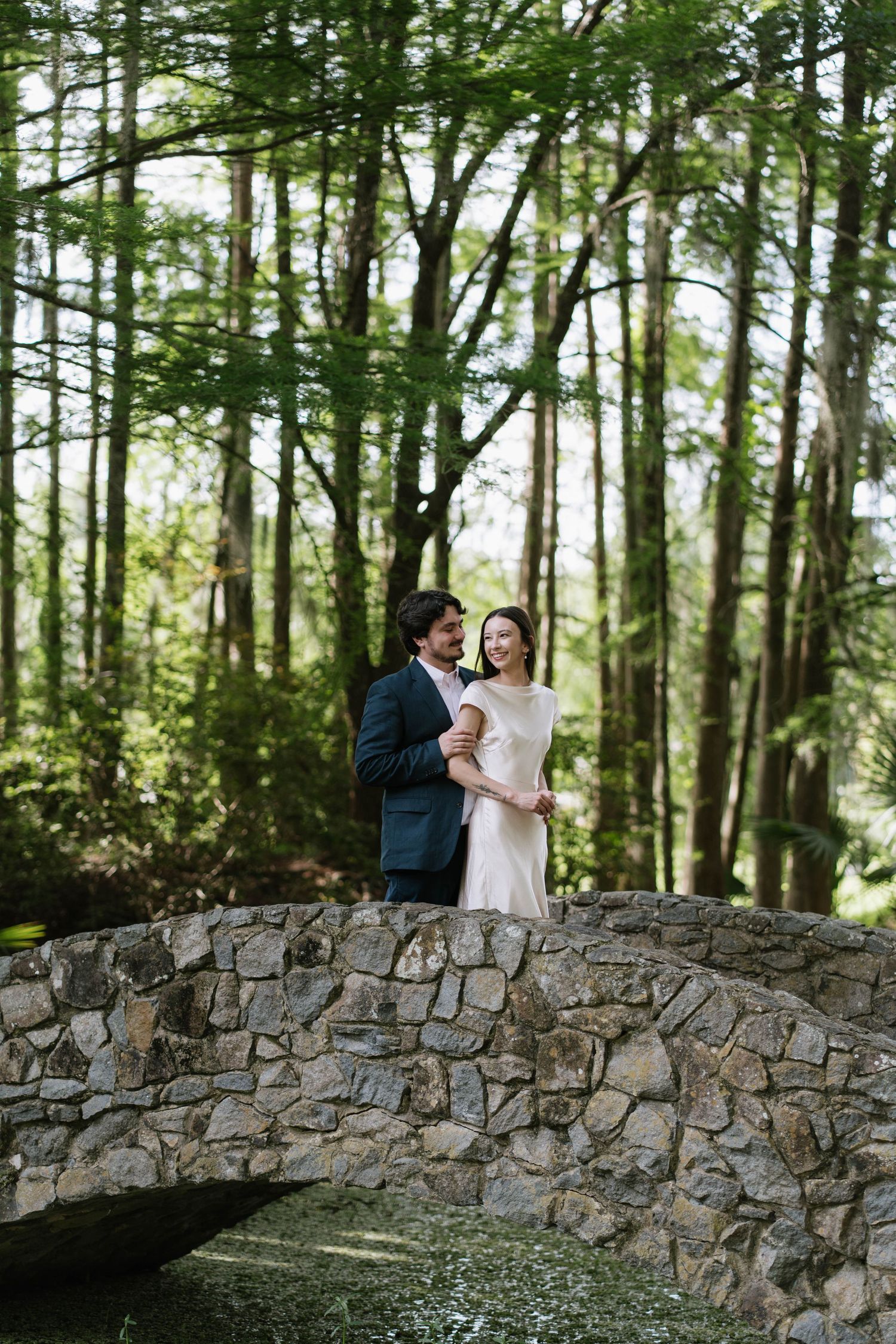 Couple embracing on a stone bridge surrounded by lush green forest trees in a romantic outdoor wedding portrait.