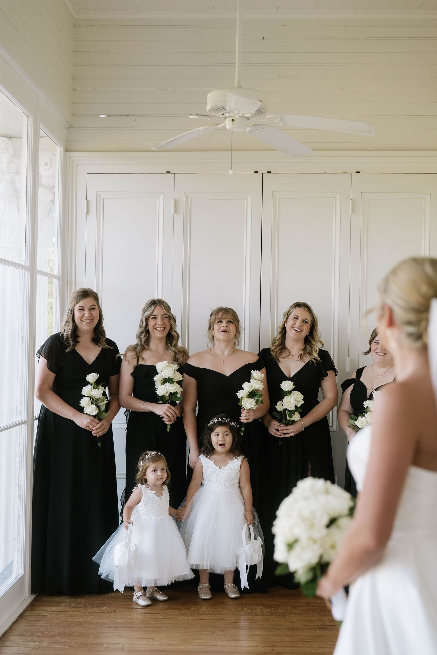 Bridesmaids in black dresses and flower girls in white tutus pose in a bright indoor space.