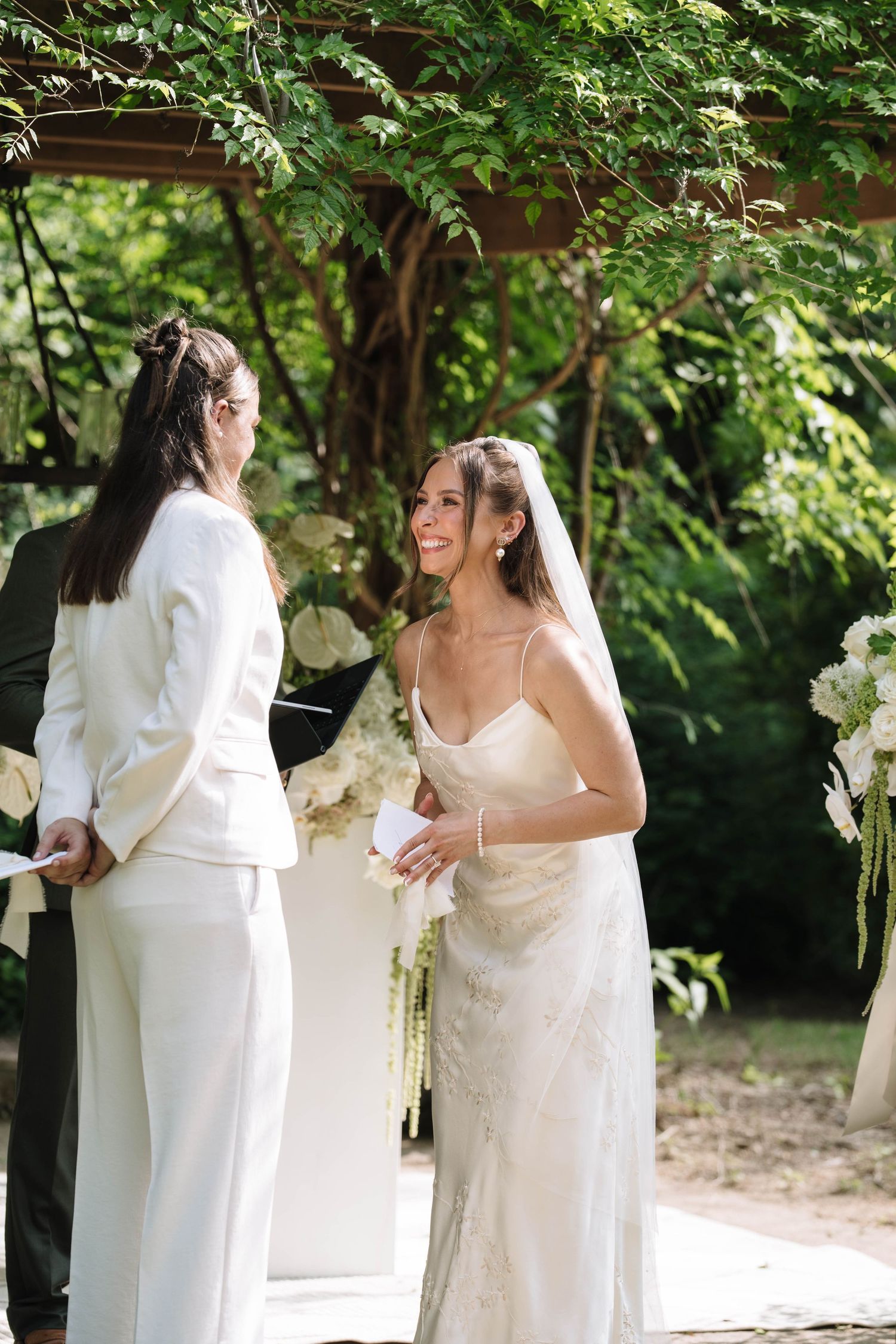 Two brides exchange vows outdoors under lush green trees, one in a white suit and one in a lace wedding gown with veil.