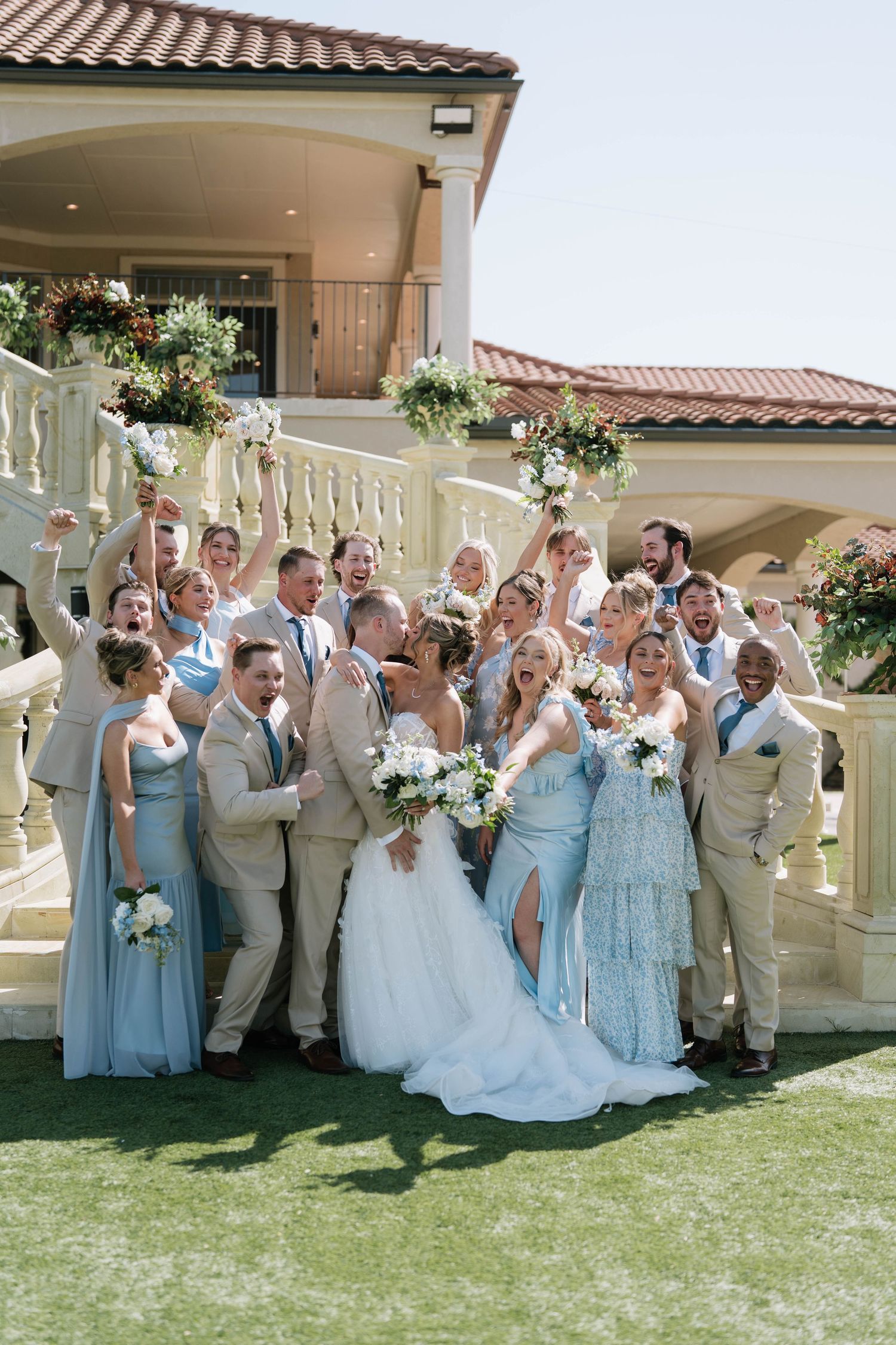 Wedding party in light blue dresses and beige suits poses enthusiastically for group photos.