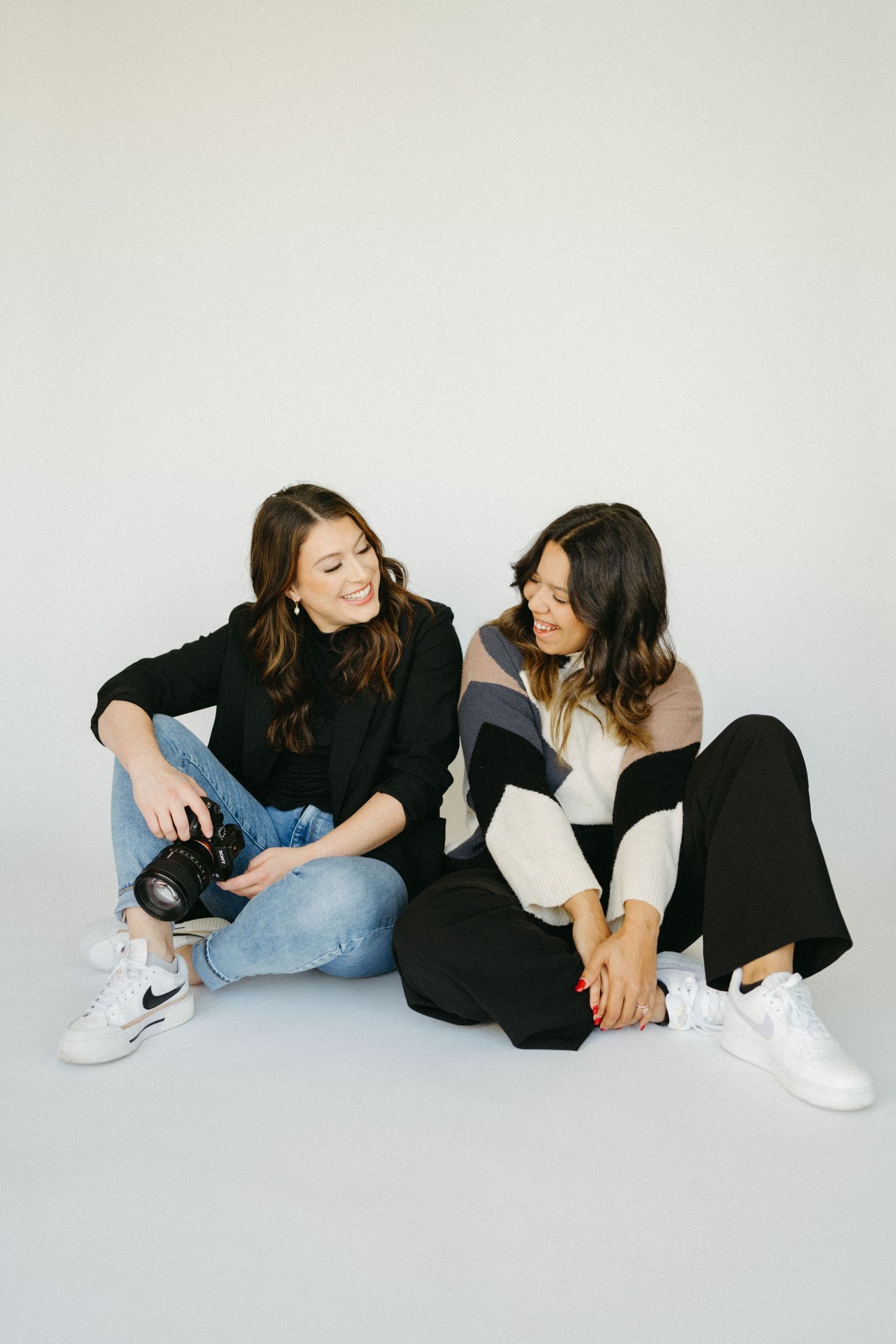 Two people in casual outfits lounging on floor during artistic photoshoot with minimalist white background.