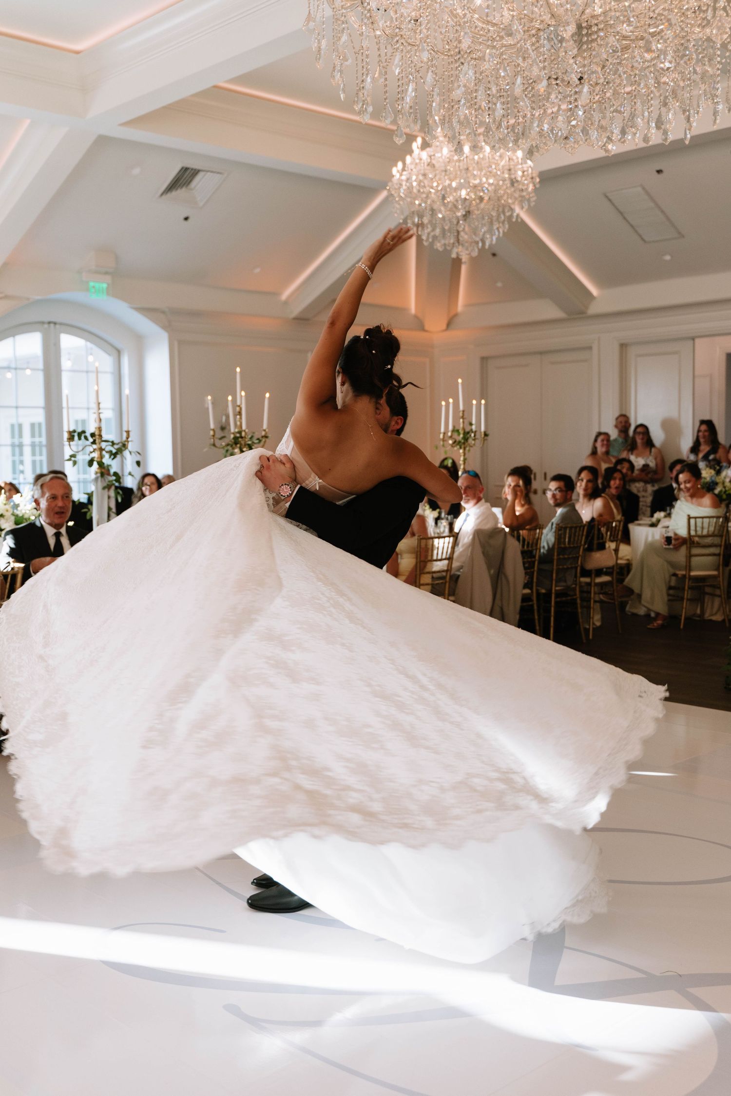 Bride and groom share first dance as wedding gown swirls elegantly in a grand ballroom with crystal chandeliers.