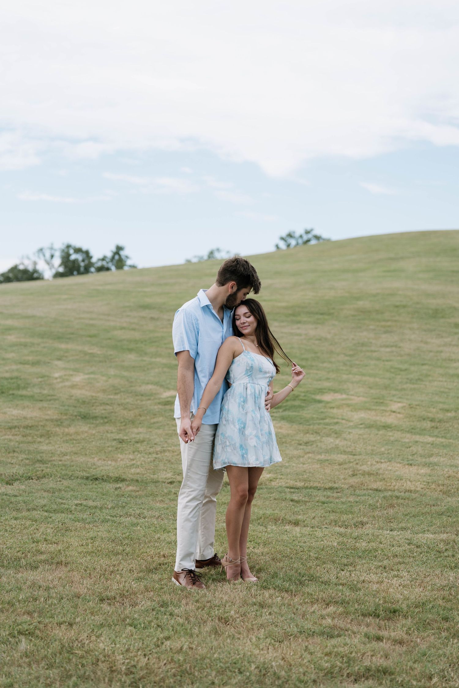 Couple in casual blue and white outfits pose playfully on grassy hillside.