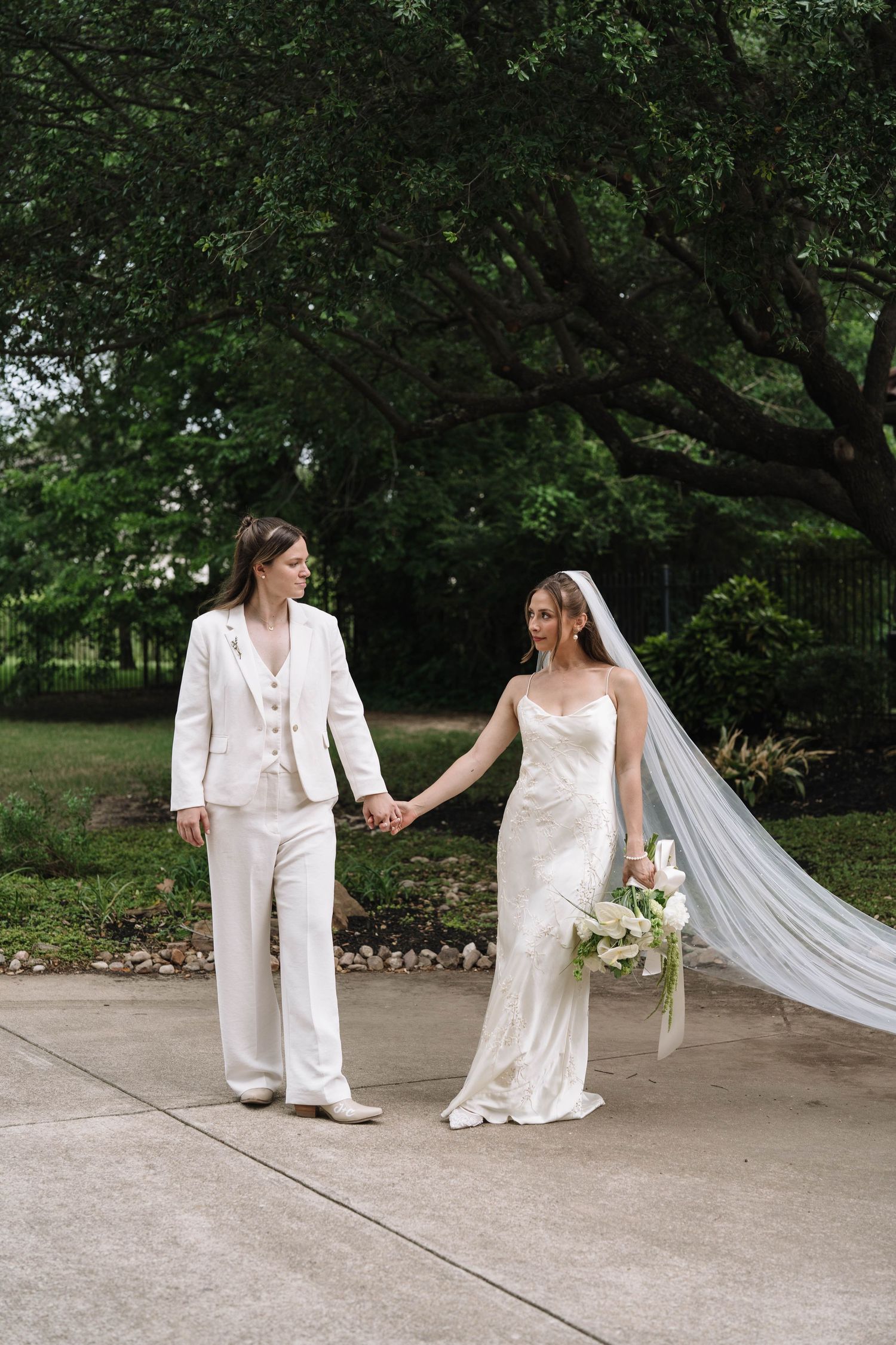Two brides hold hands outdoors, one in a white suit and boots, the other in a satin gown with veil and bouquet.