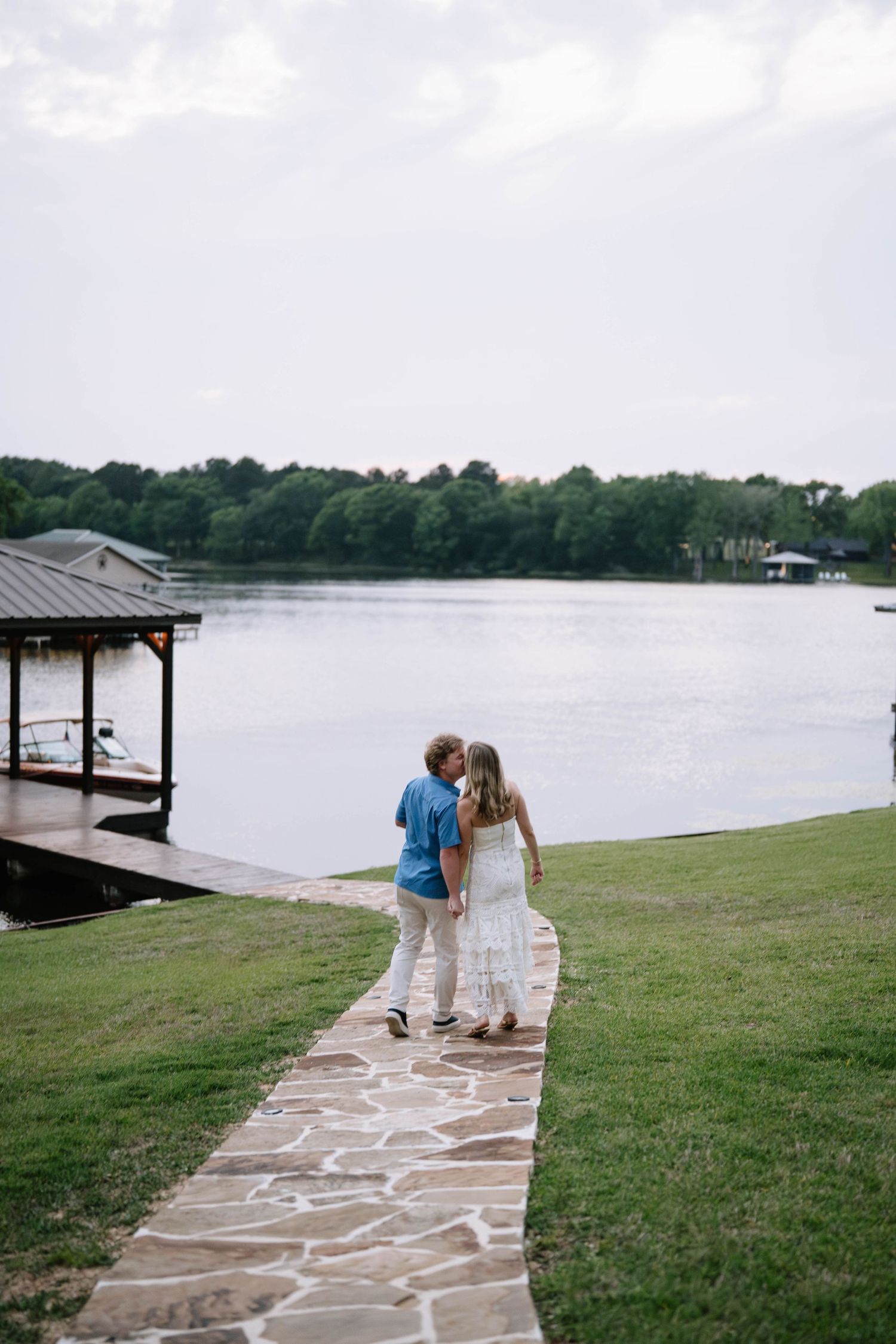 Couple walking on stone path by lake near boat dock, woman in white dress, man in blue shirt, lush green trees.