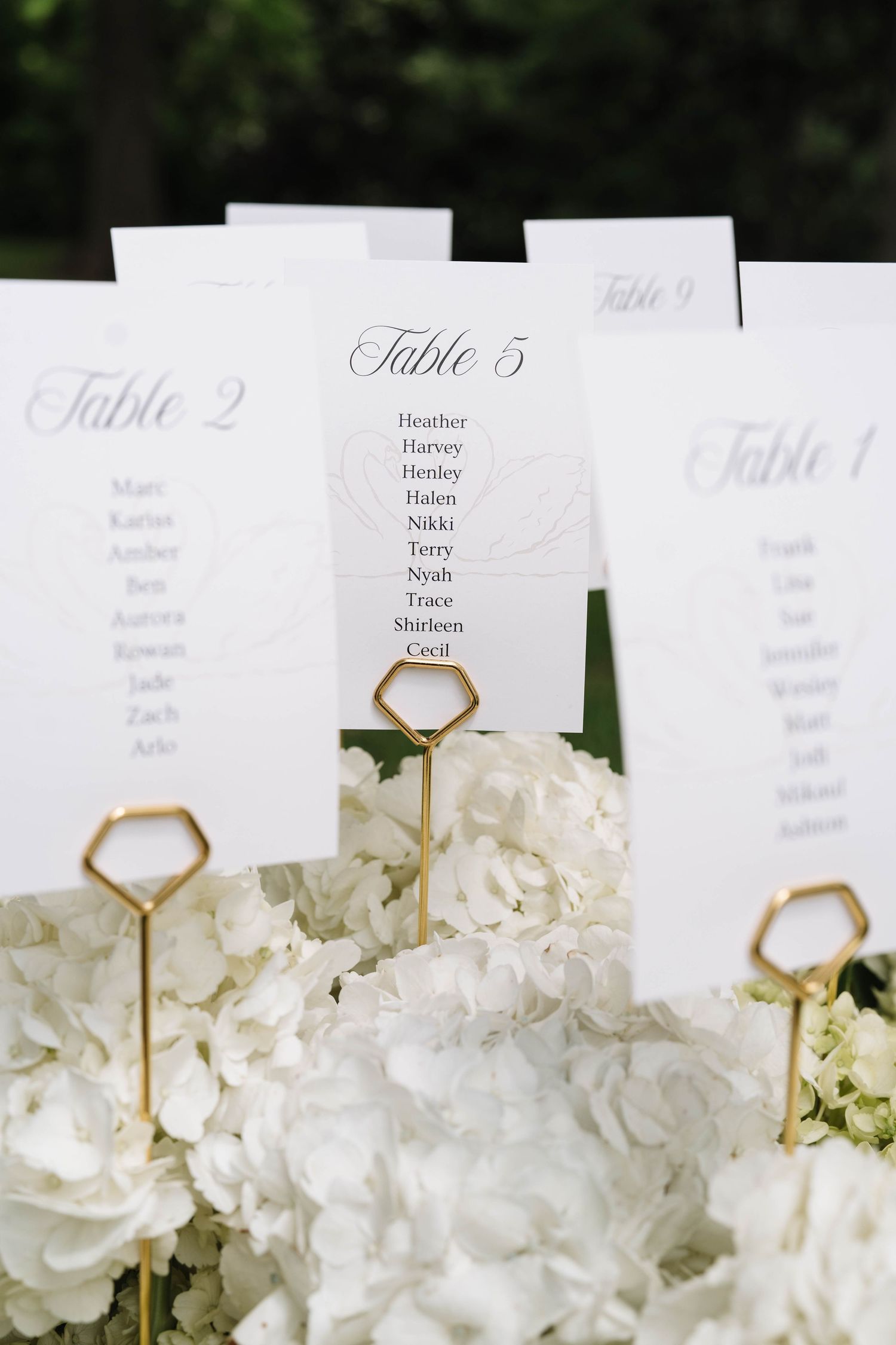 Elegant wedding seating chart cards in gold geometric holders surrounded by white hydrangeas.