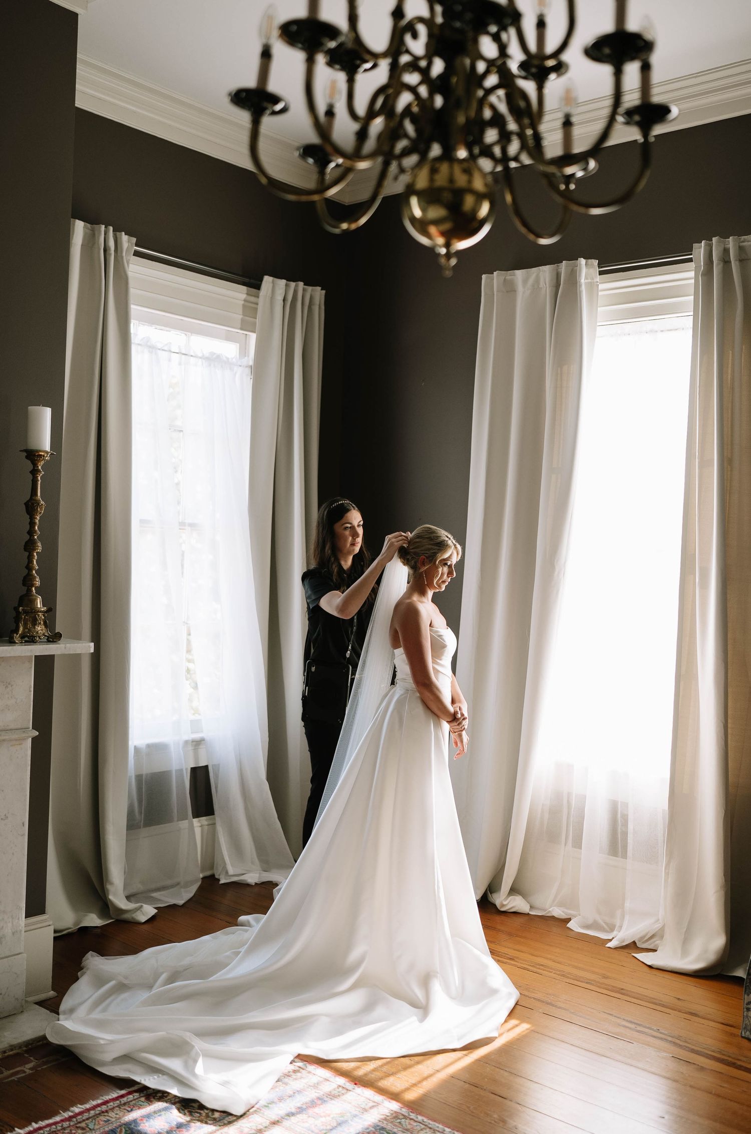 Final wedding dress preparations in an elegant room with white curtains and a dramatic chandelier.