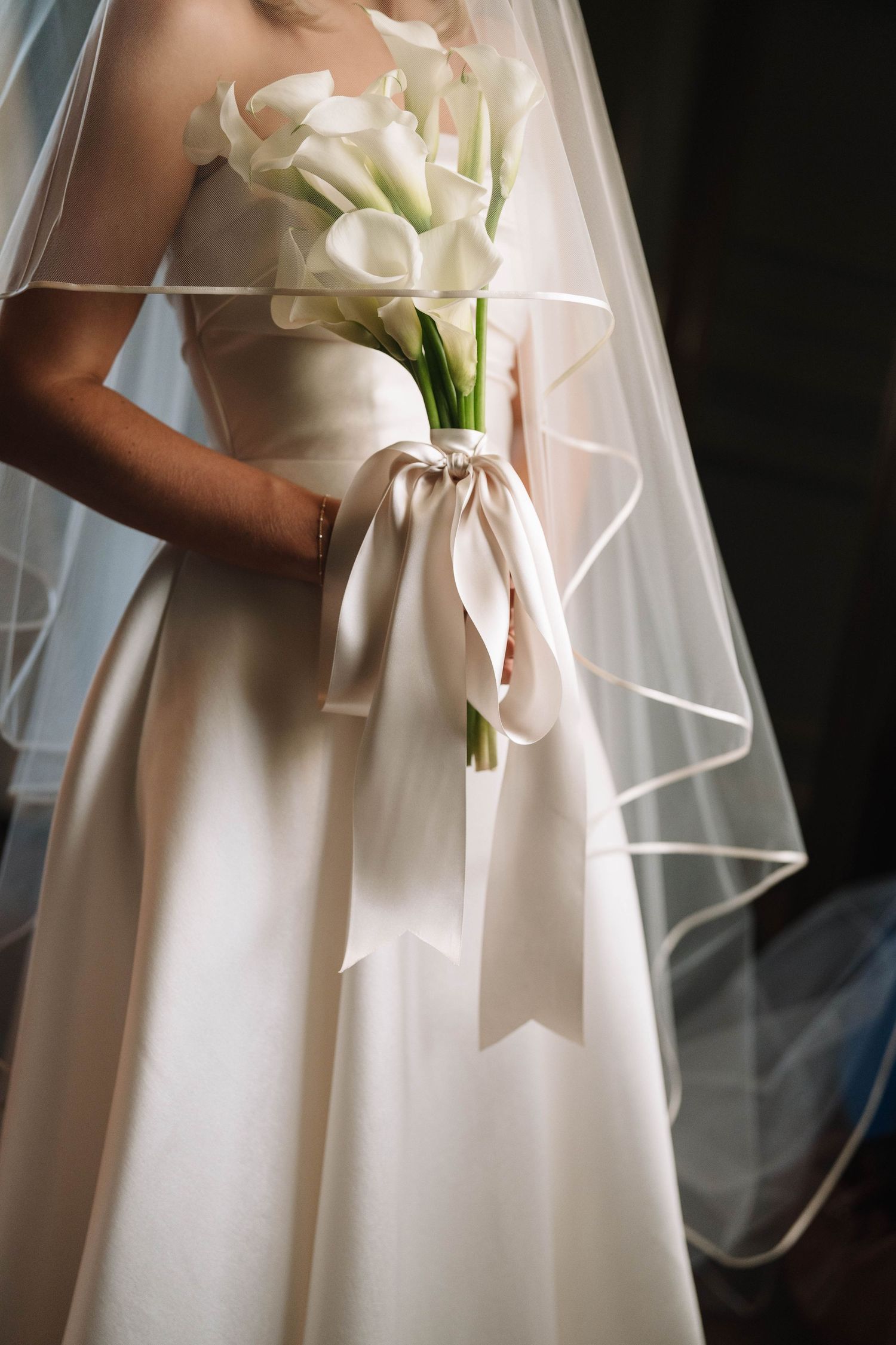 Bride in white satin gown holding calla lily bouquet with ribbon, wearing veil.