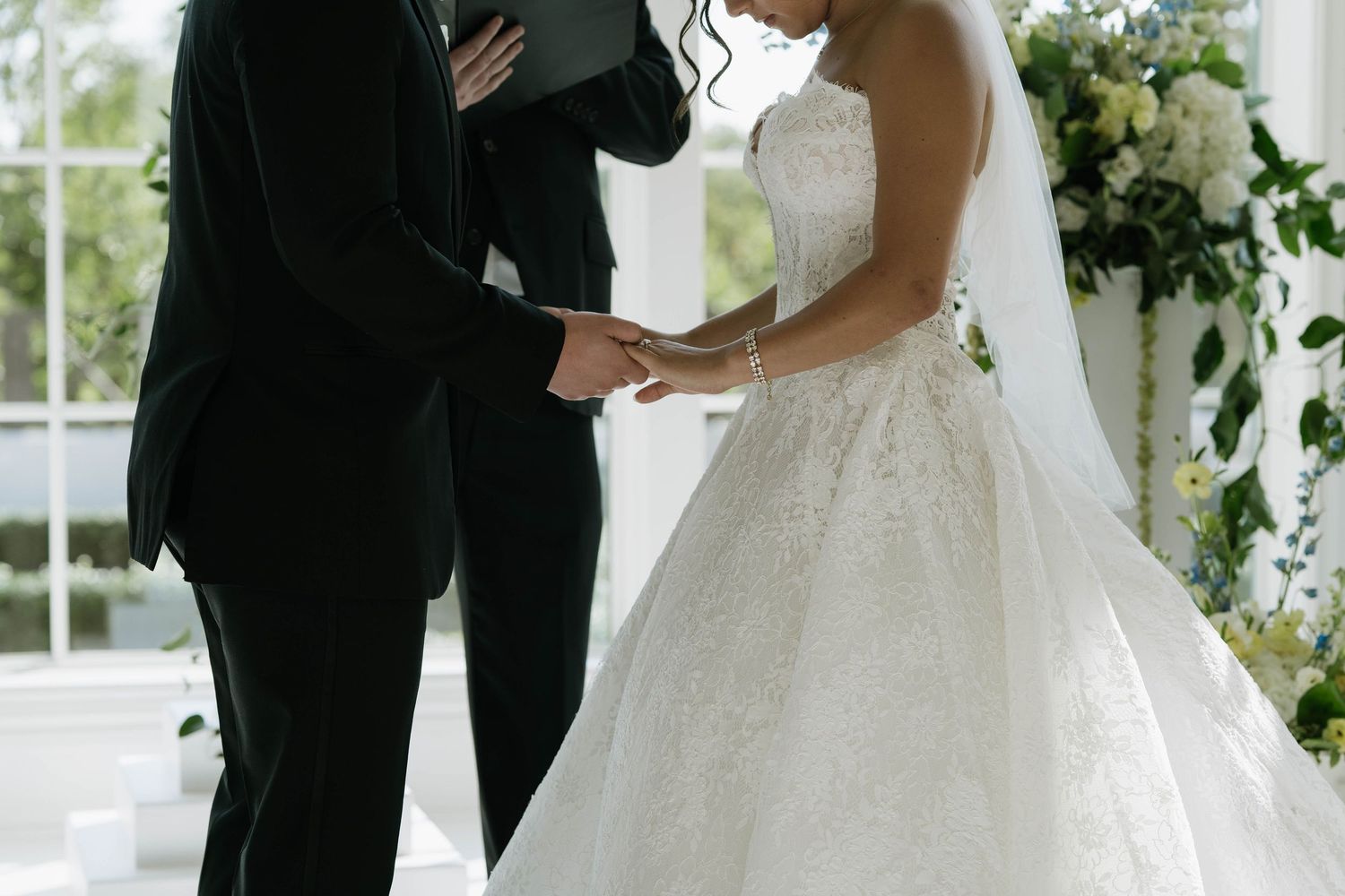 Bride in lace ball gown and groom in black suit exchange vows at elegant wedding ceremony with floral backdrop.