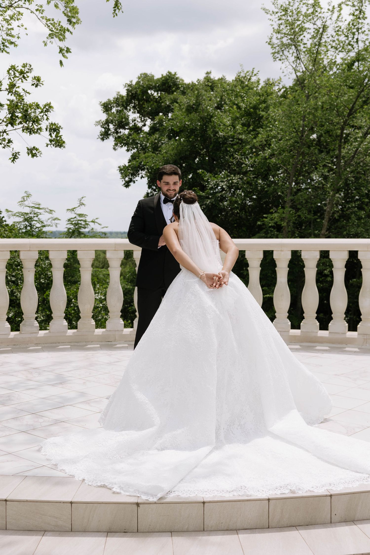 Bride in elegant white gown with veil and groom in black tuxedo embrace on a white balcony surrounded by lush trees.