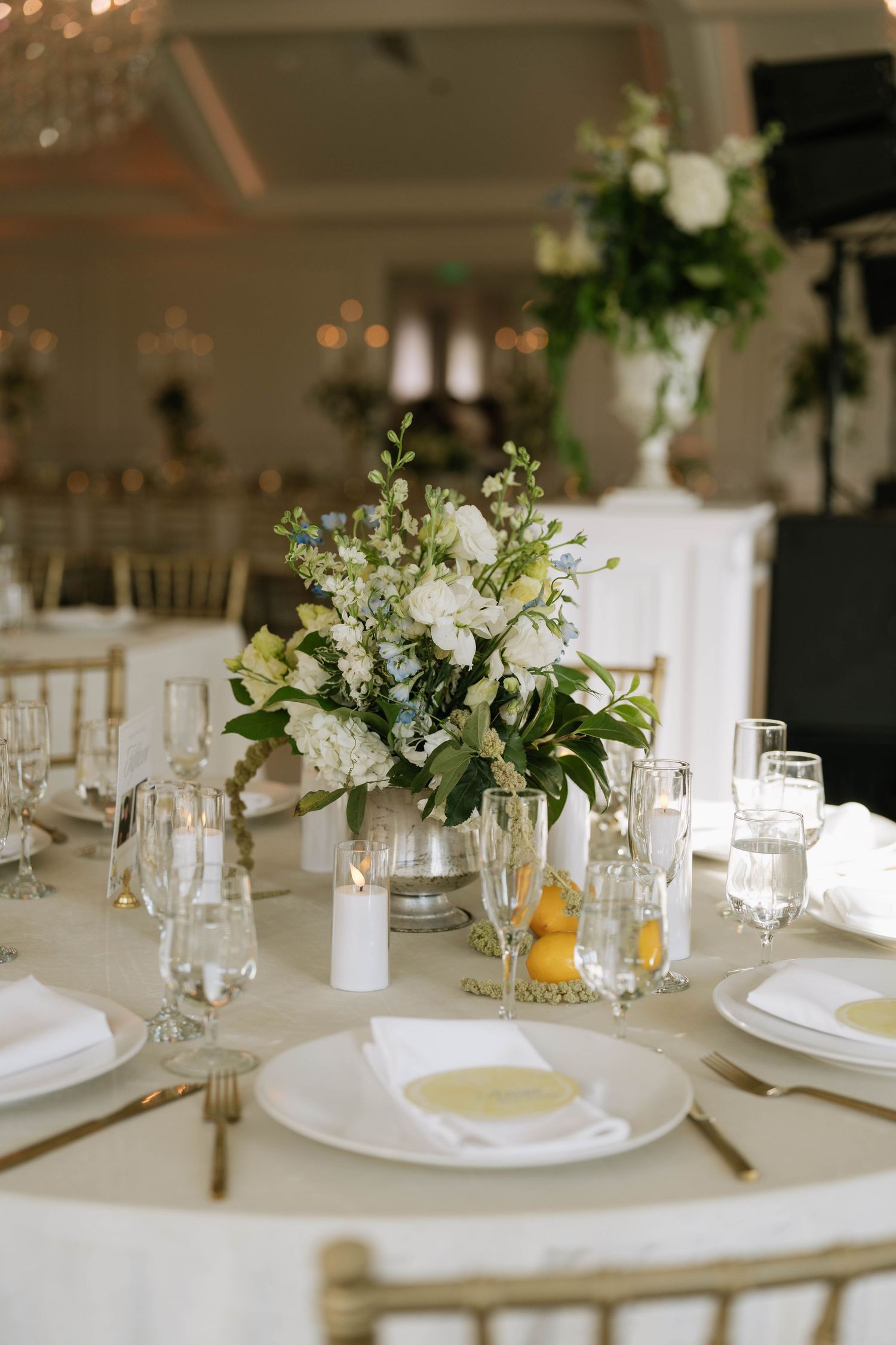Elegant wedding reception table with white floral centerpiece, gold chiavari chairs, crystal glassware, and white linen napkins.