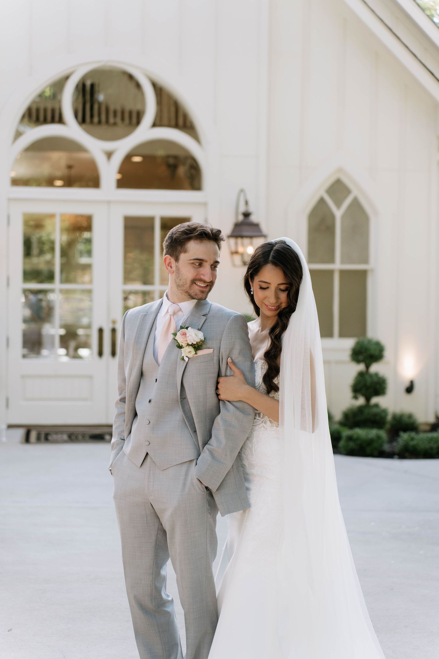 Newlyweds in grey suit and white dress walking outside their white chapel venue.