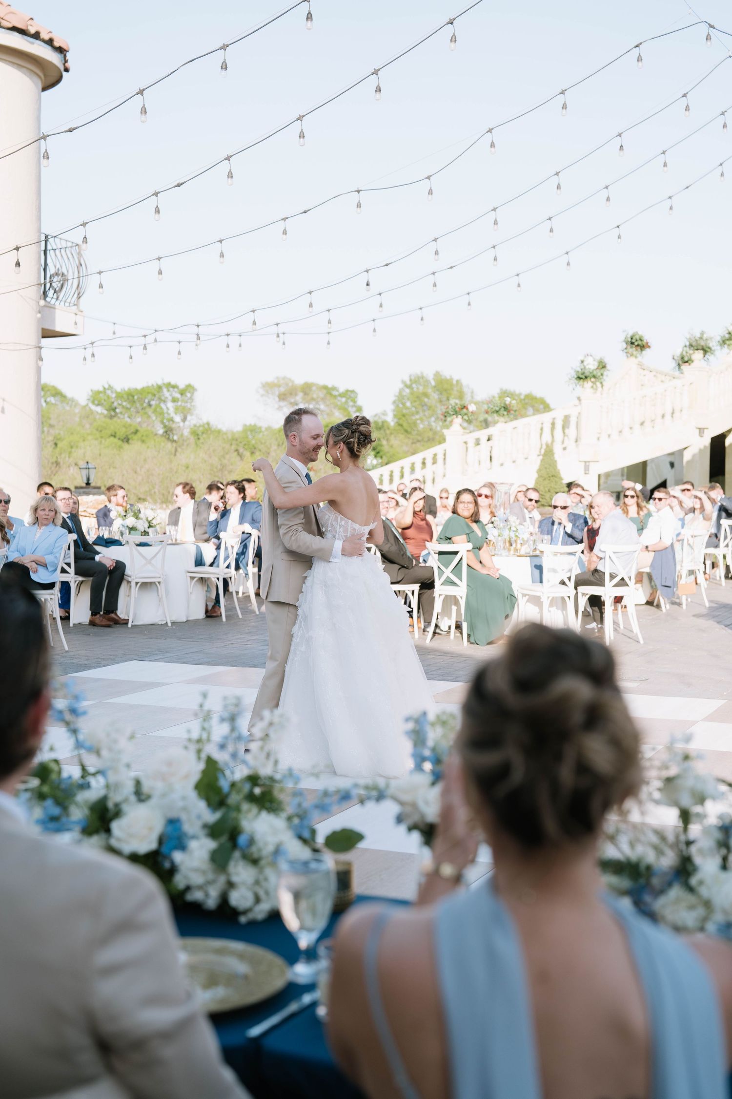An outdoor wedding reception setup with string lights and white tables under a covered patio.