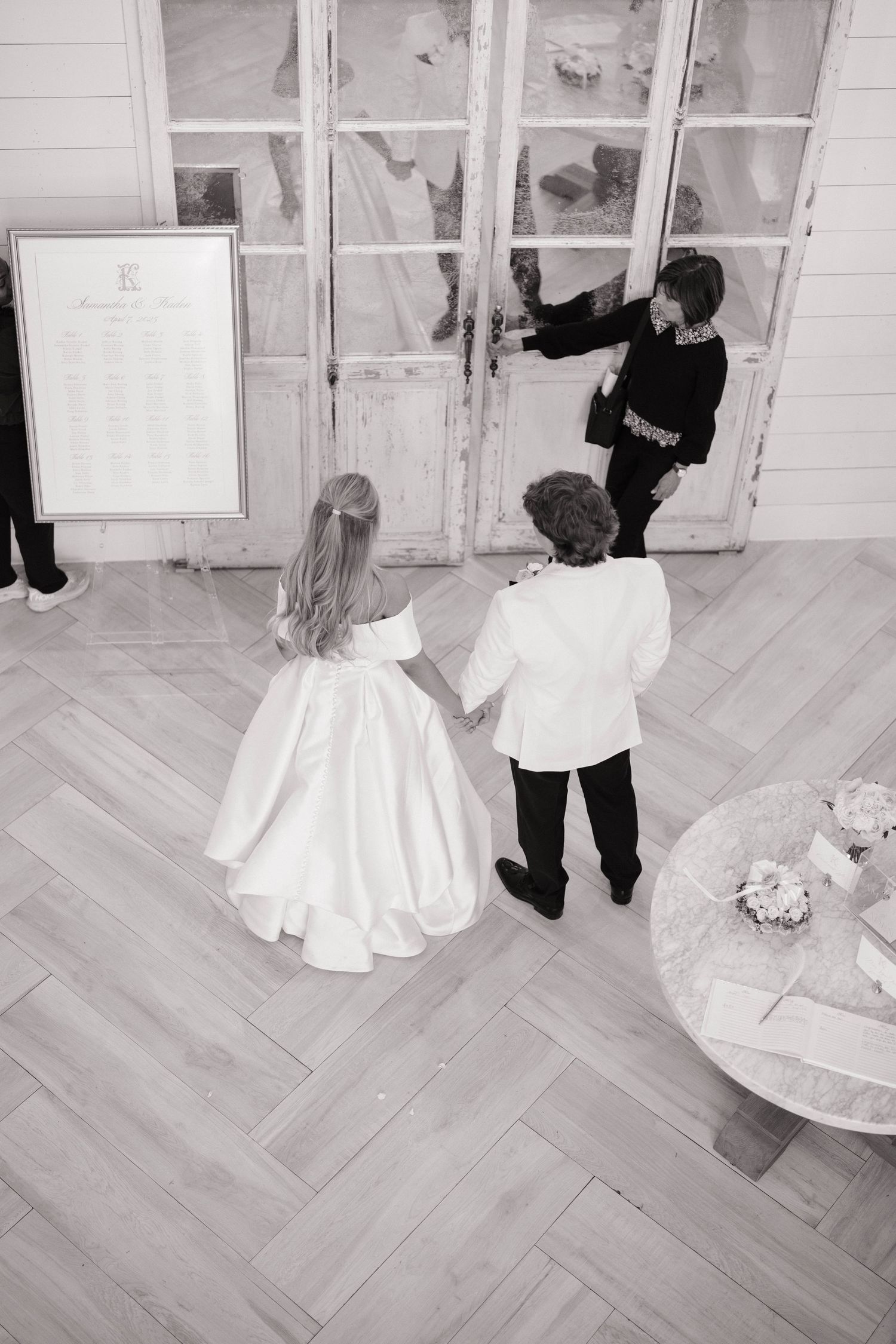 Two children in formal wear peek through glass doors at a wedding venue.