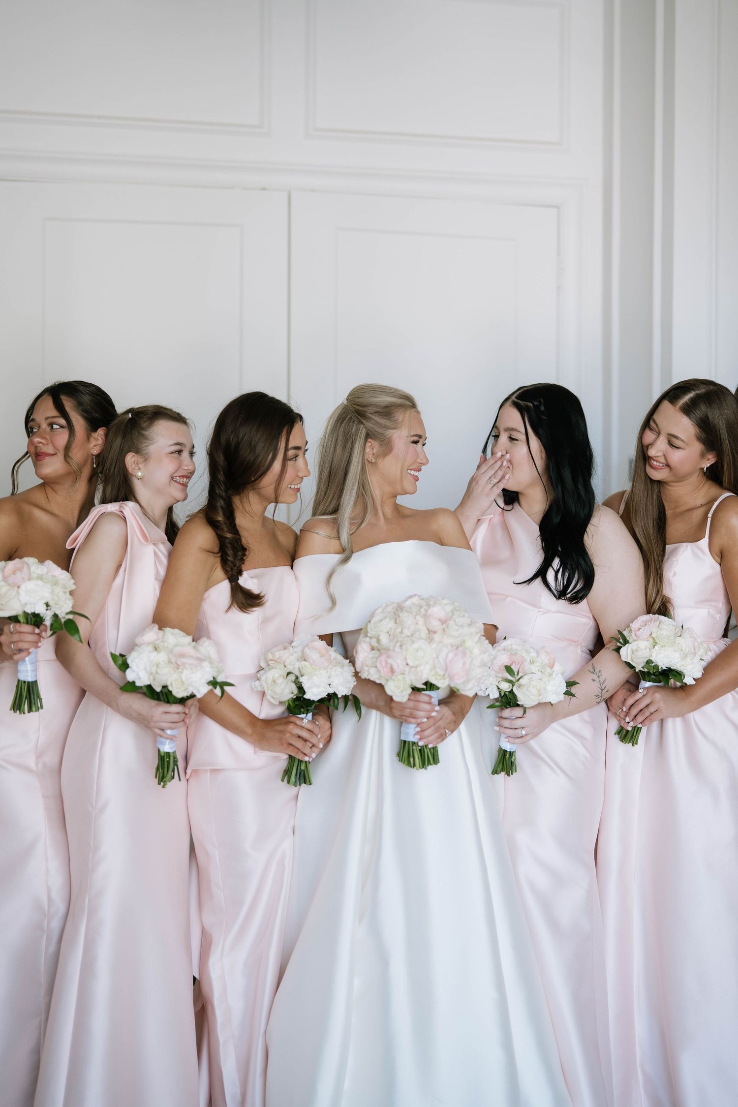 Bridesmaids in blush pink dresses lined up against white wall holding white bouquets.