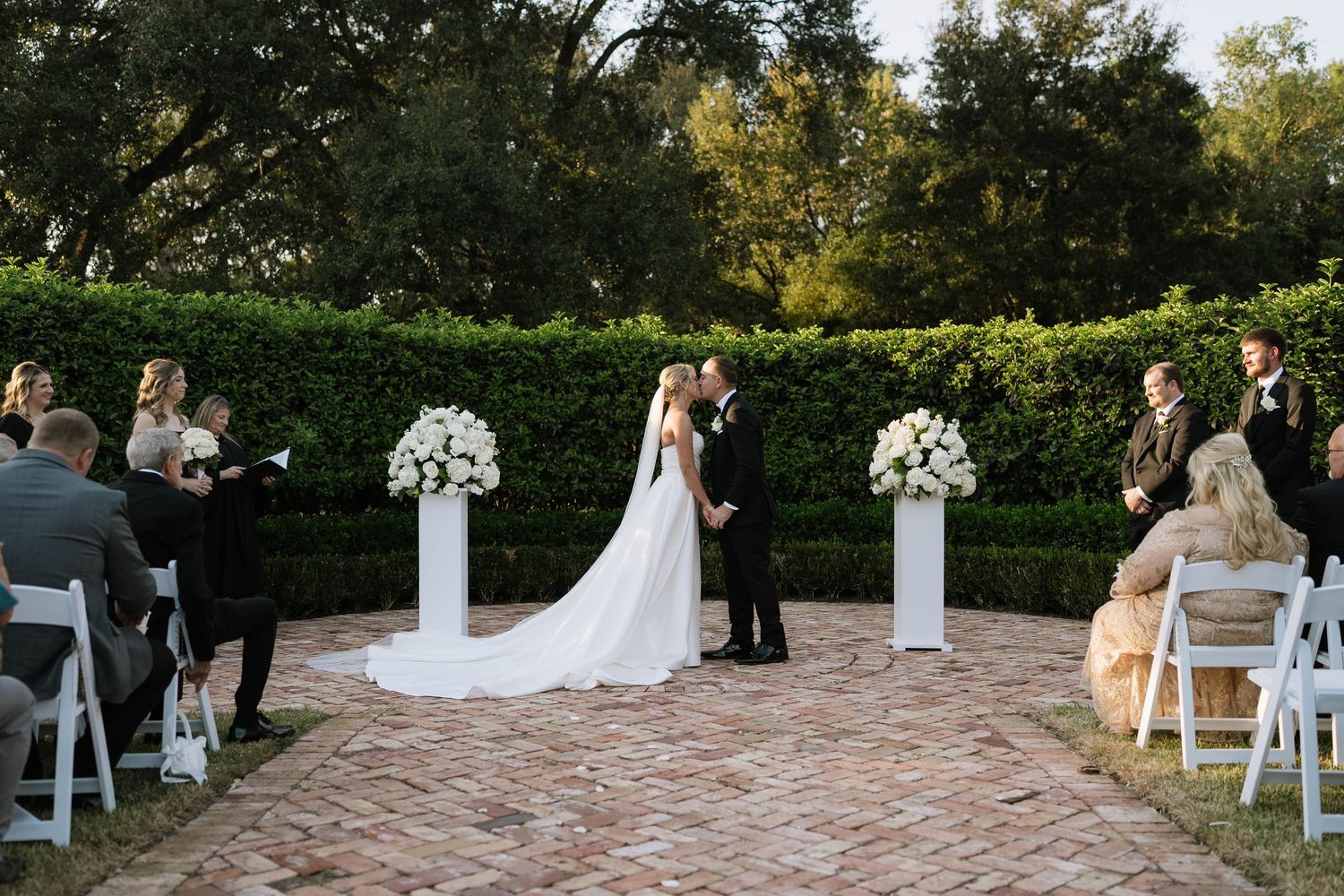 Outdoor wedding ceremony setup with white chairs and floral arrangements on a stone patio.