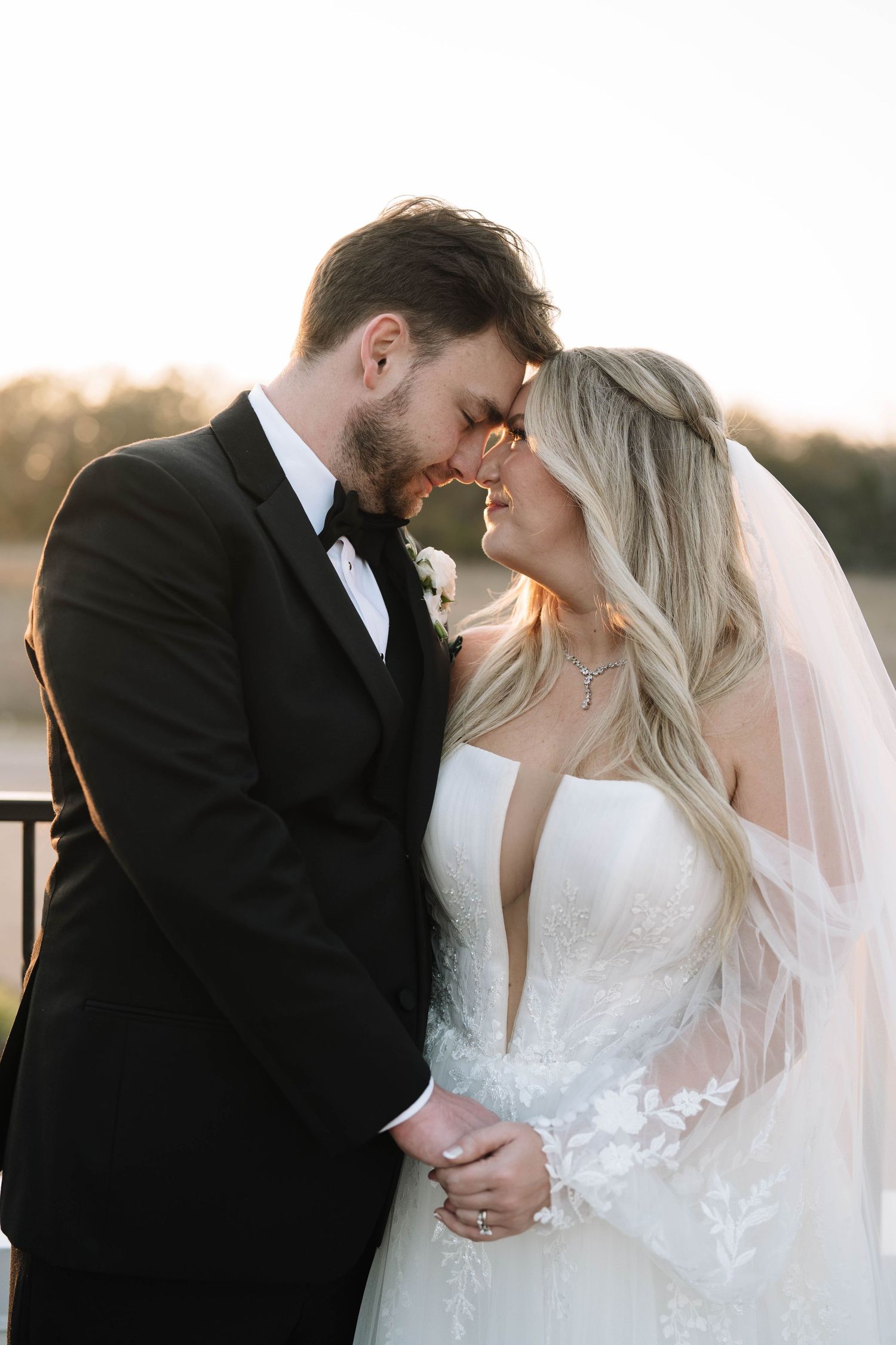 Bride and groom touching foreheads tenderly in golden sunset light, she in lace gown with veil, he in black tuxedo.