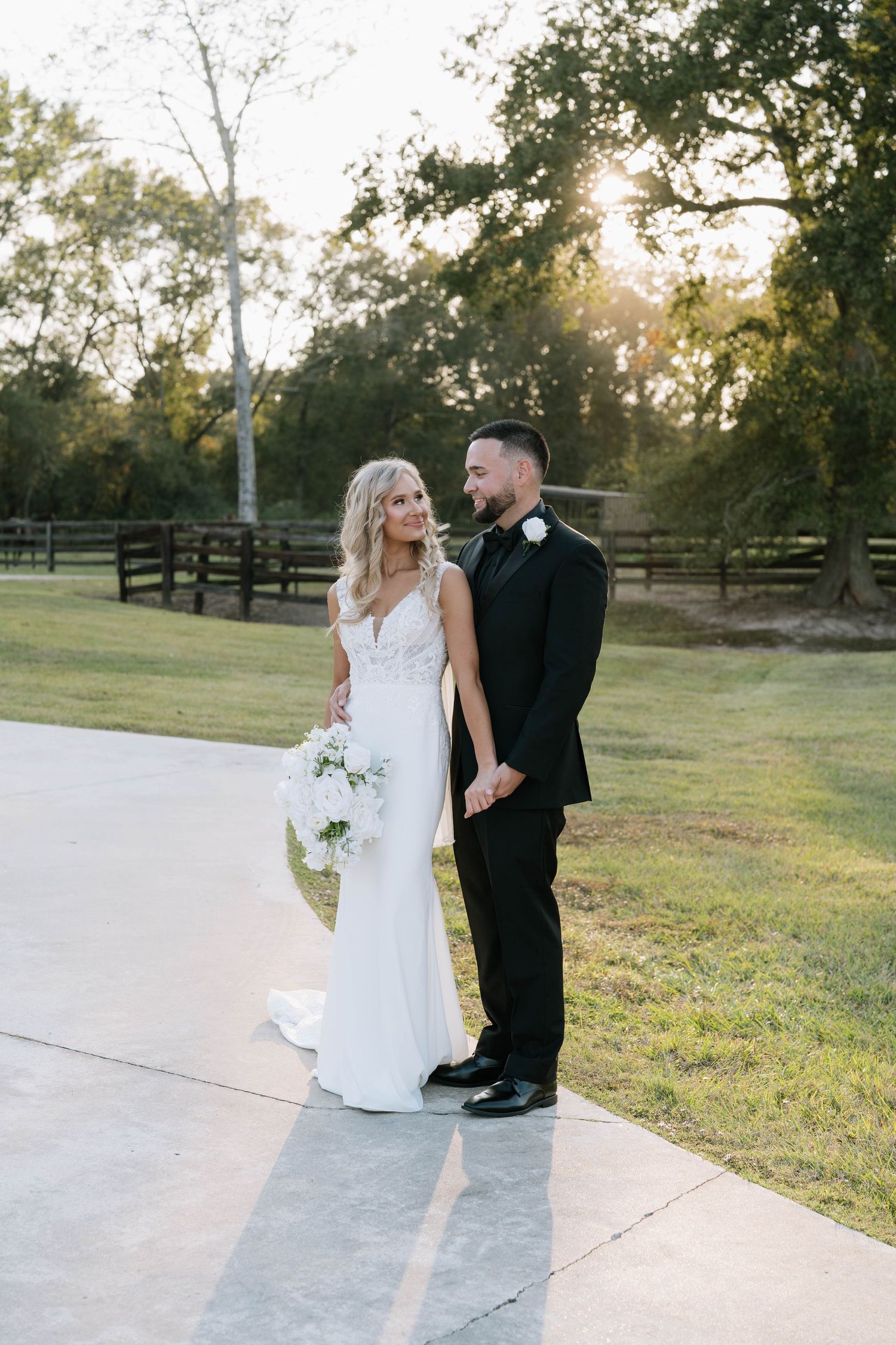 Wedding couple walking together on a paved pathway surrounded by green grass and trees at sunset.
