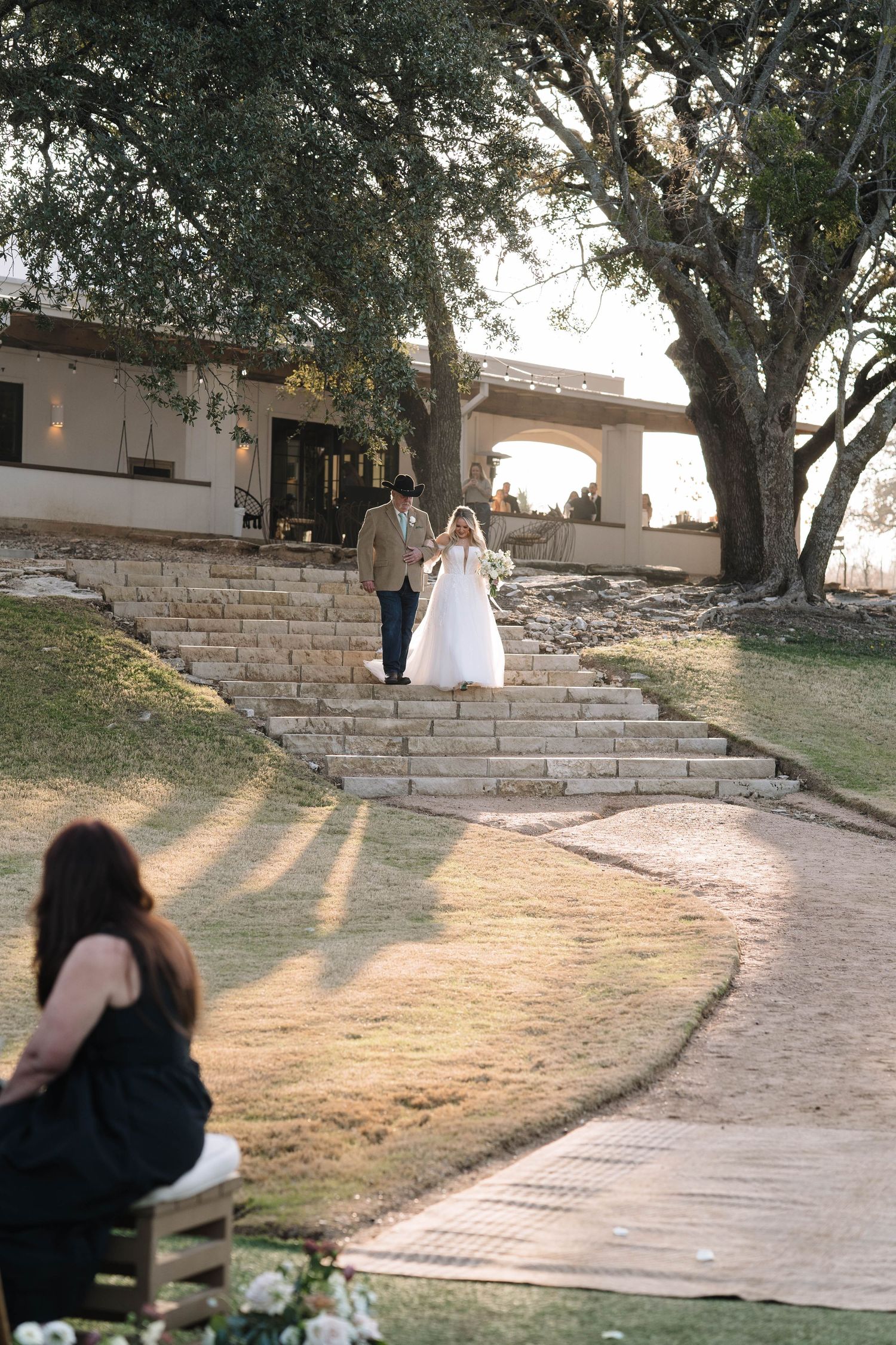 Bride and groom walk down stone steps at an outdoor wedding venue surrounded by large oak trees.