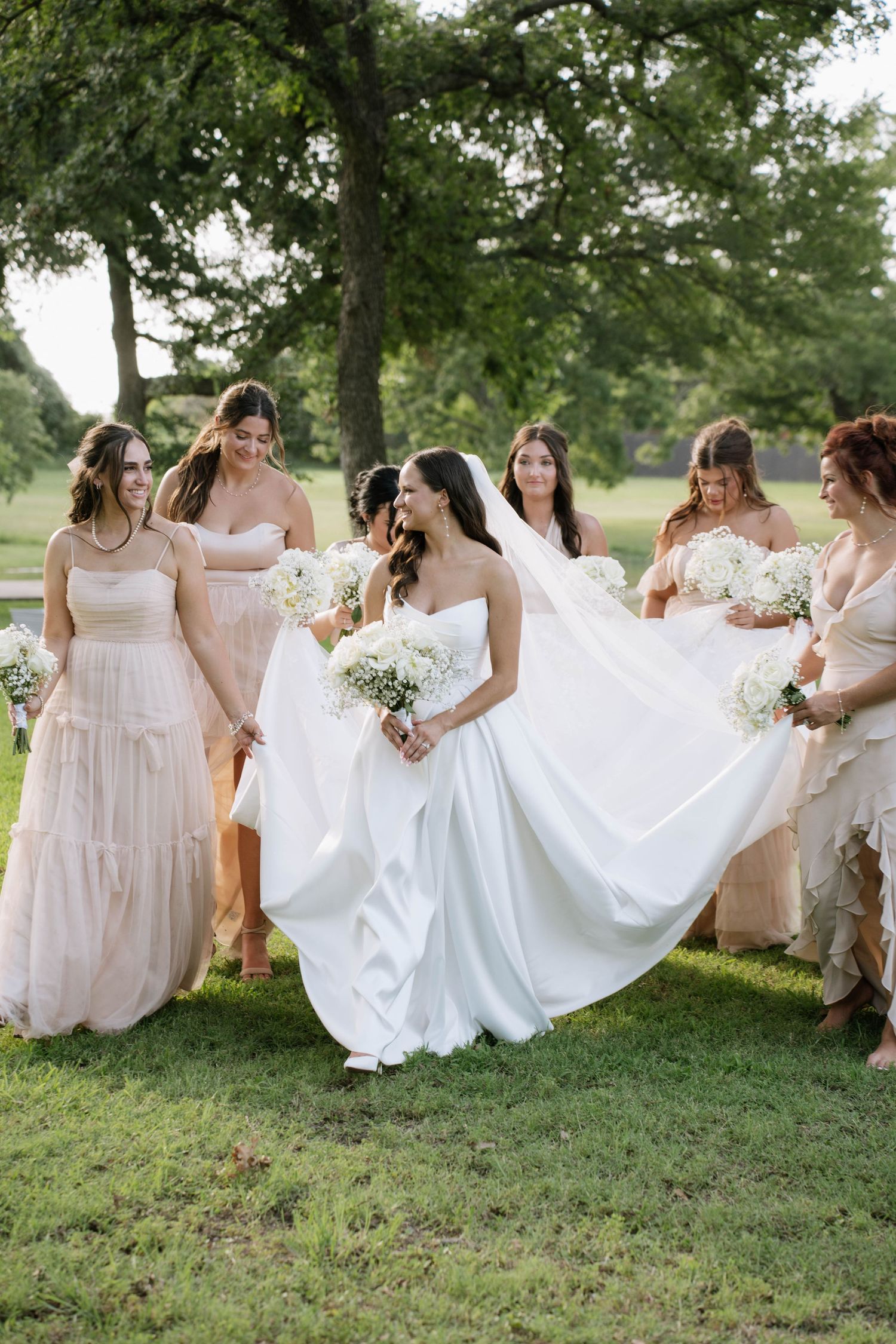Bridesmaids in blush dresses help adjust the bride's flowing white gown on a grassy lawn under trees.