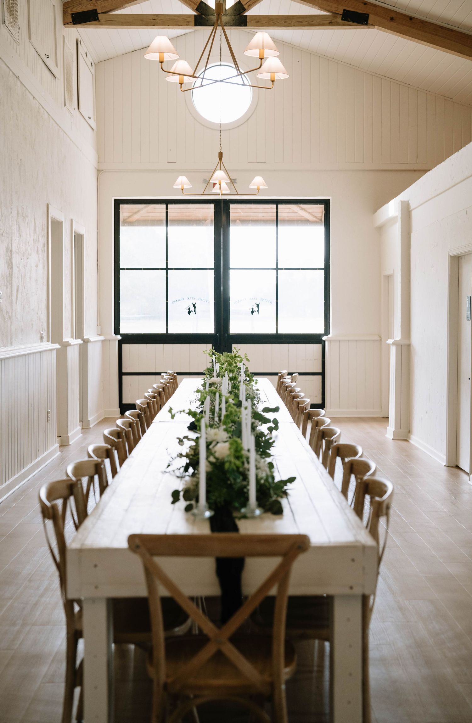 Modern white reception venue with long wooden dining tables and vaulted ceiling with chandeliers.