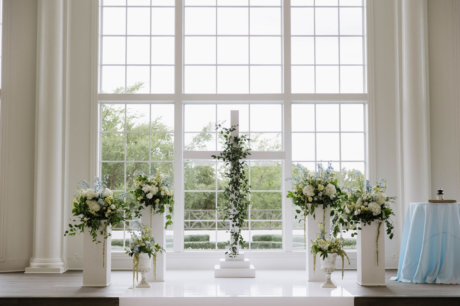 Elegant wedding altar with white floral arrangements and a cross centerpiece in front of large windows.