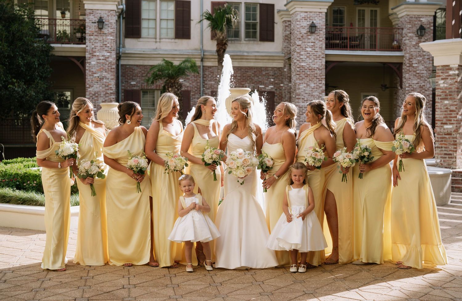 Bridesmaids in pale yellow dresses pose with the bride in white outside a stone building.