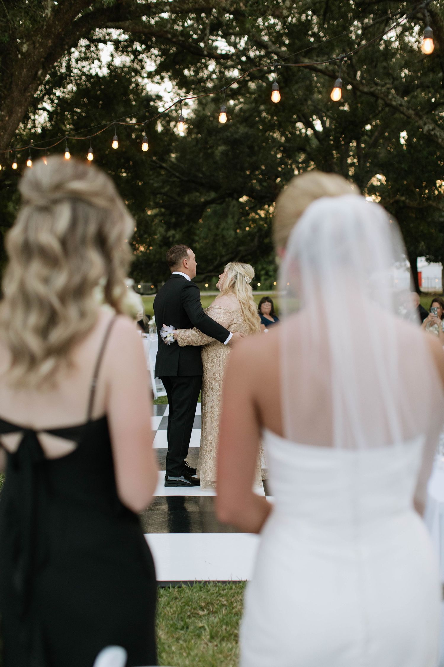 Wedding ceremony setup outdoors with string lights hanging from trees as guests gather to watch.