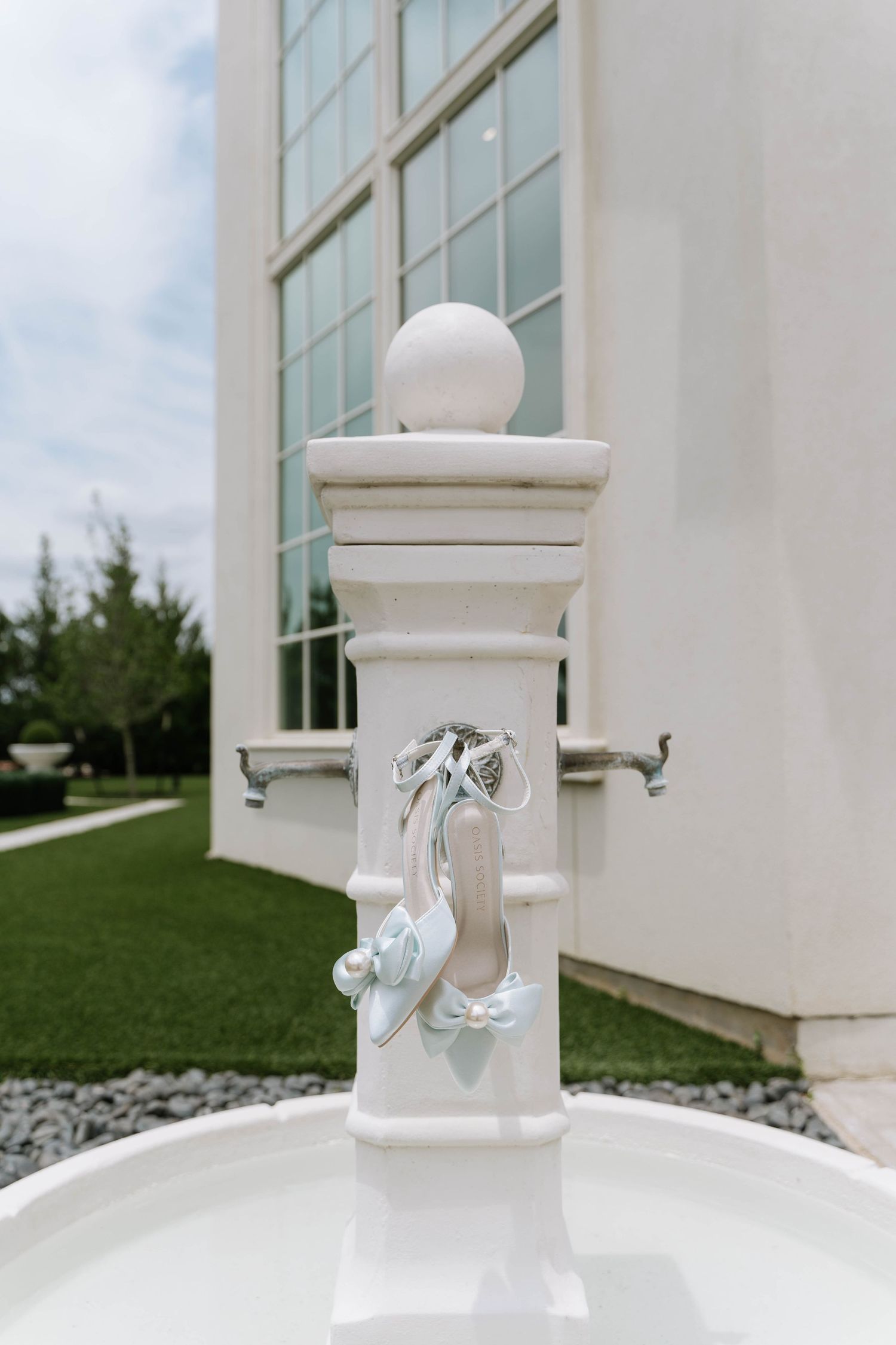 Elegant white bridal heels hanging on a decorative fountain pillar outdoors at a wedding venue.