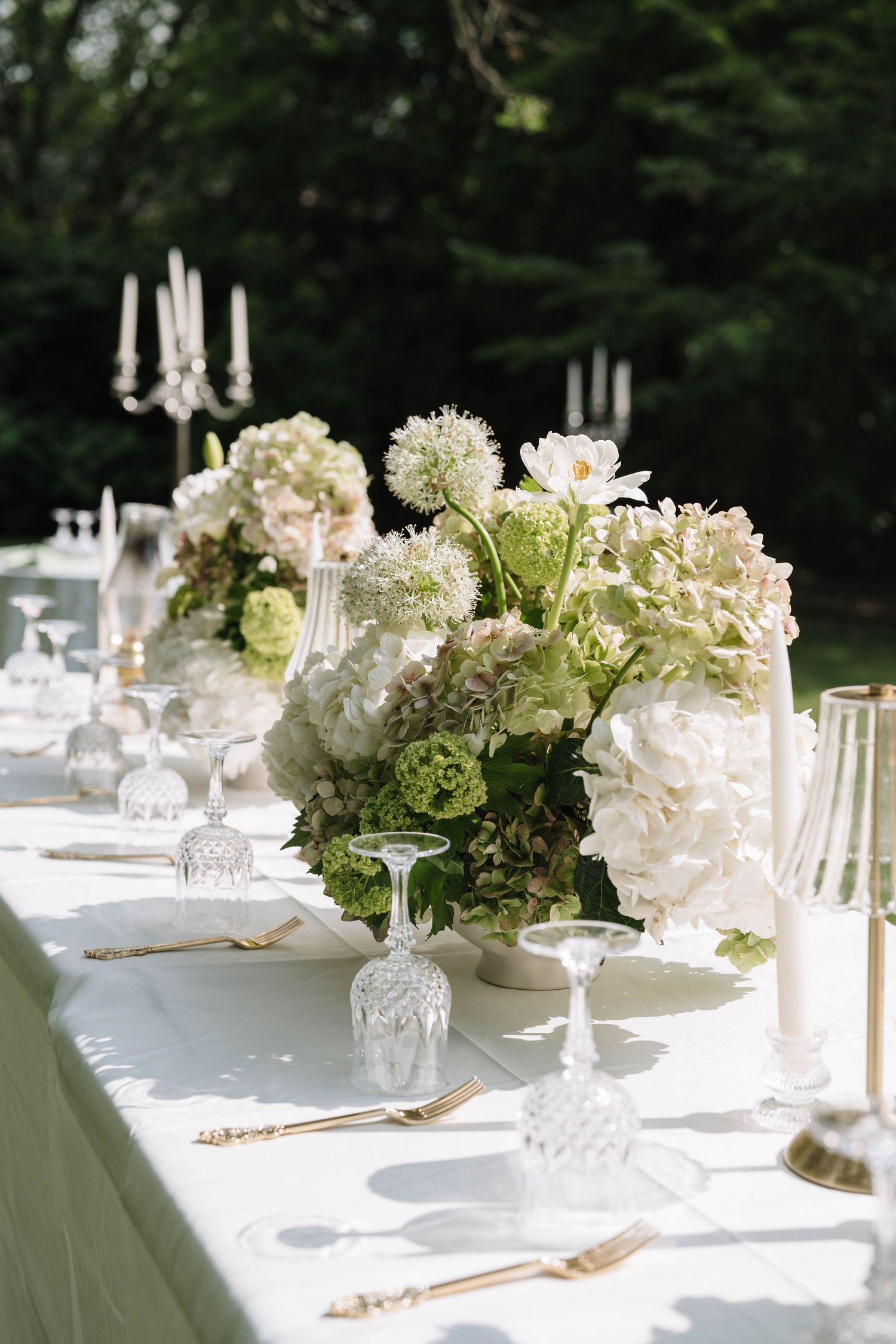 Elegant outdoor wedding reception table with white hydrangeas, crystal glasses, gold cutlery, and candelabras on a white linen.