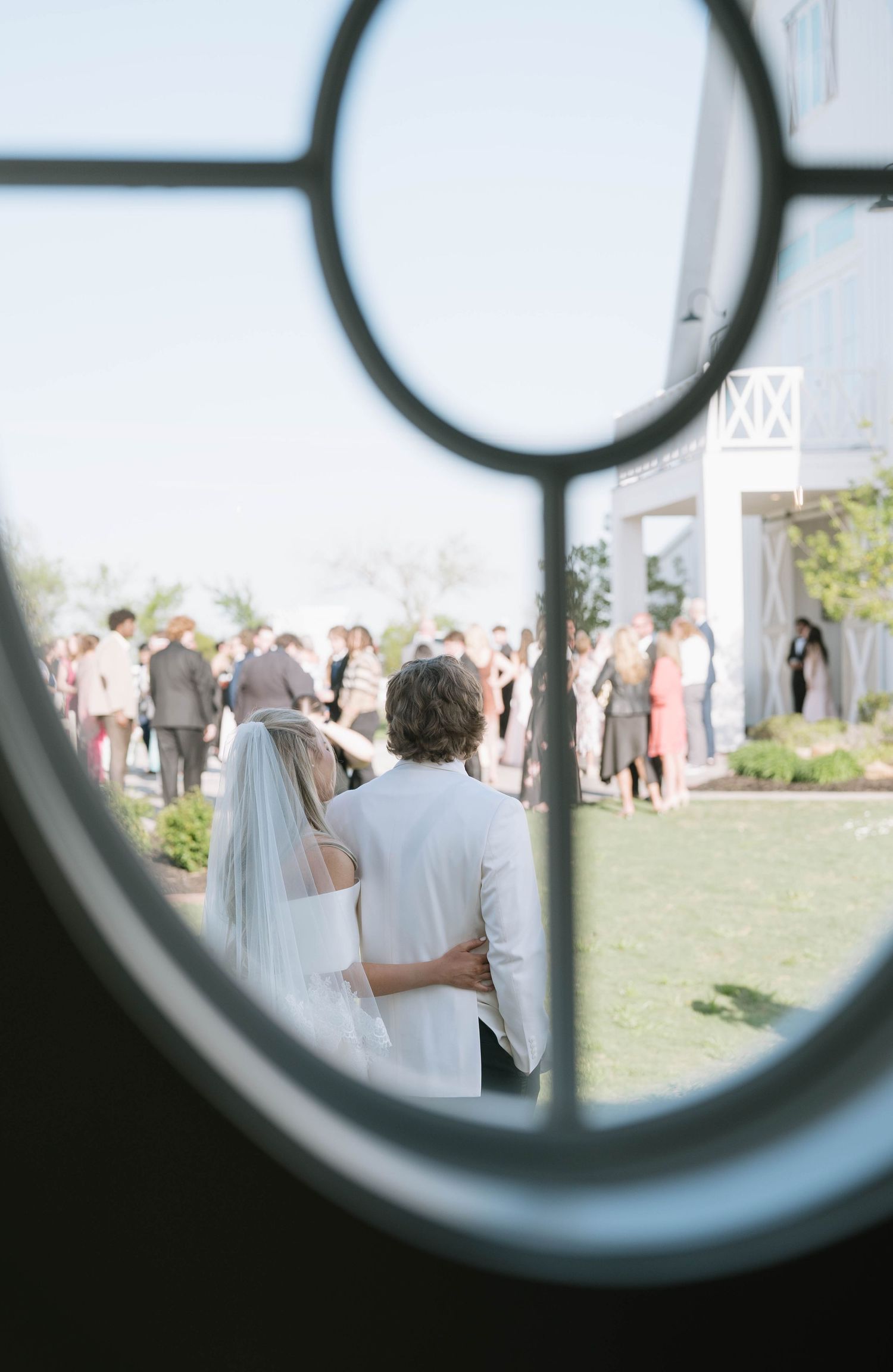 Wedding guests gather on a sunny lawn viewed through a circular window frame.