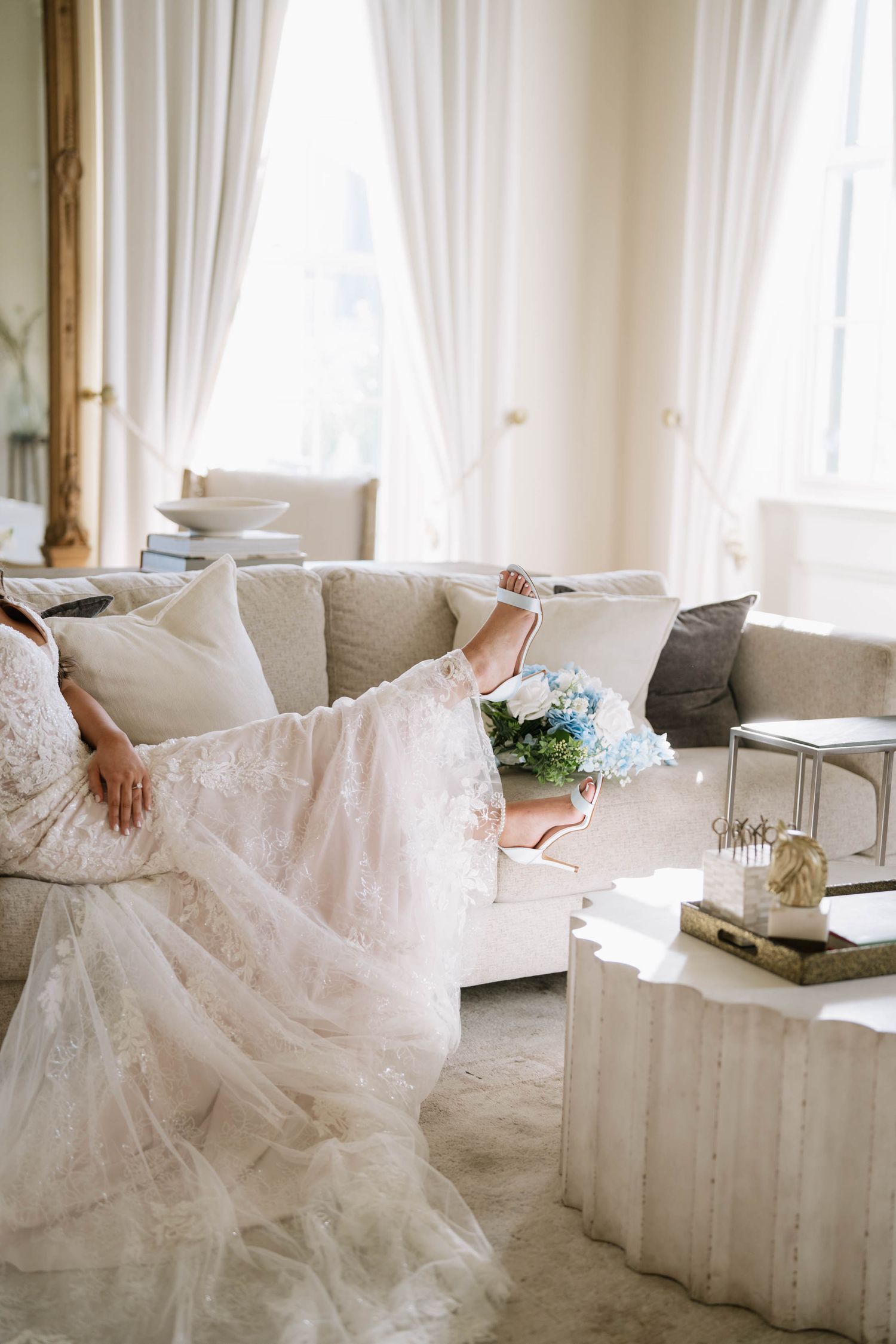 Bride in lace wedding gown relaxing on elegant cream sofa with blue floral bouquet in bright, airy room.