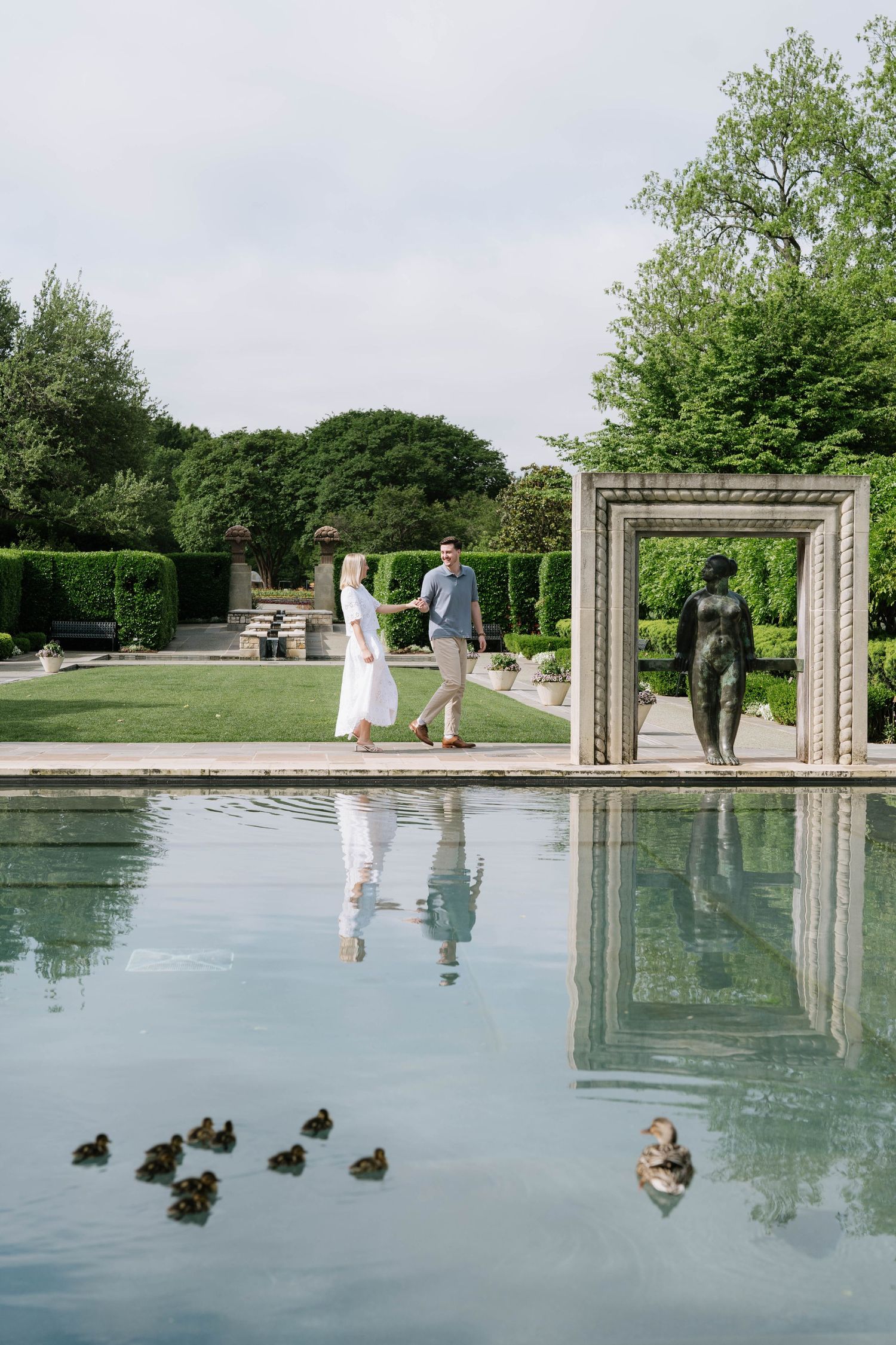 A couple walks near a reflecting pool with ducks and architectural frame in an elegant garden setting.