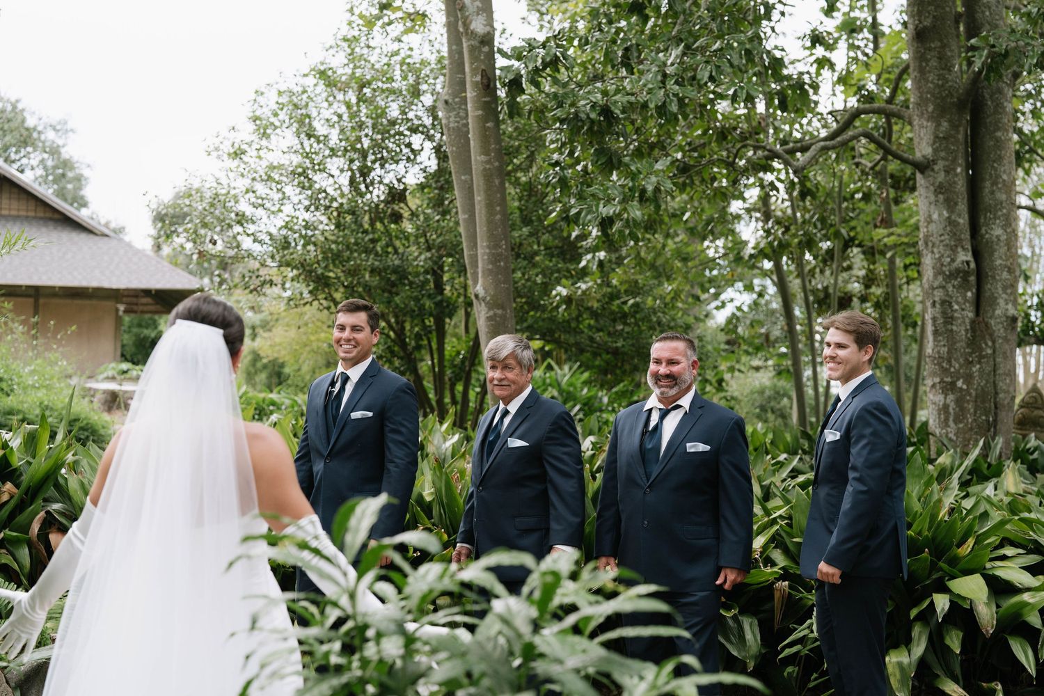 Wedding party in formal attire standing outdoors among lush garden landscaping.