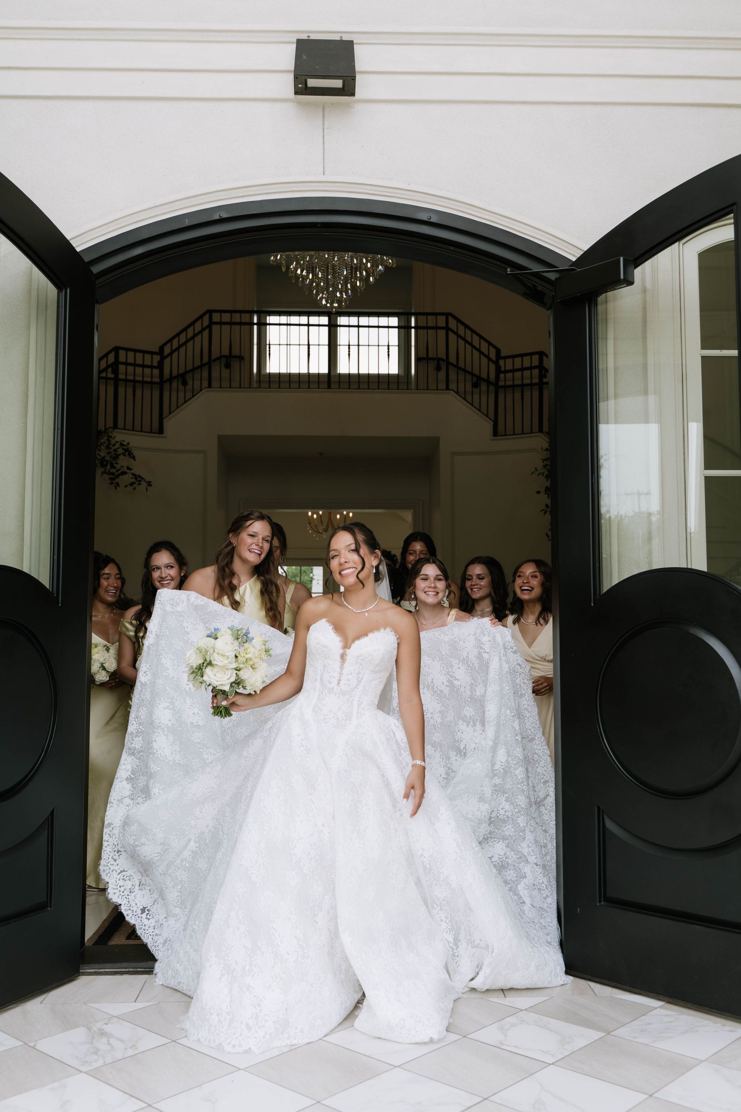 Bride exits grand double doors in white ballgown, bridesmaids holding train, holding bouquet.
