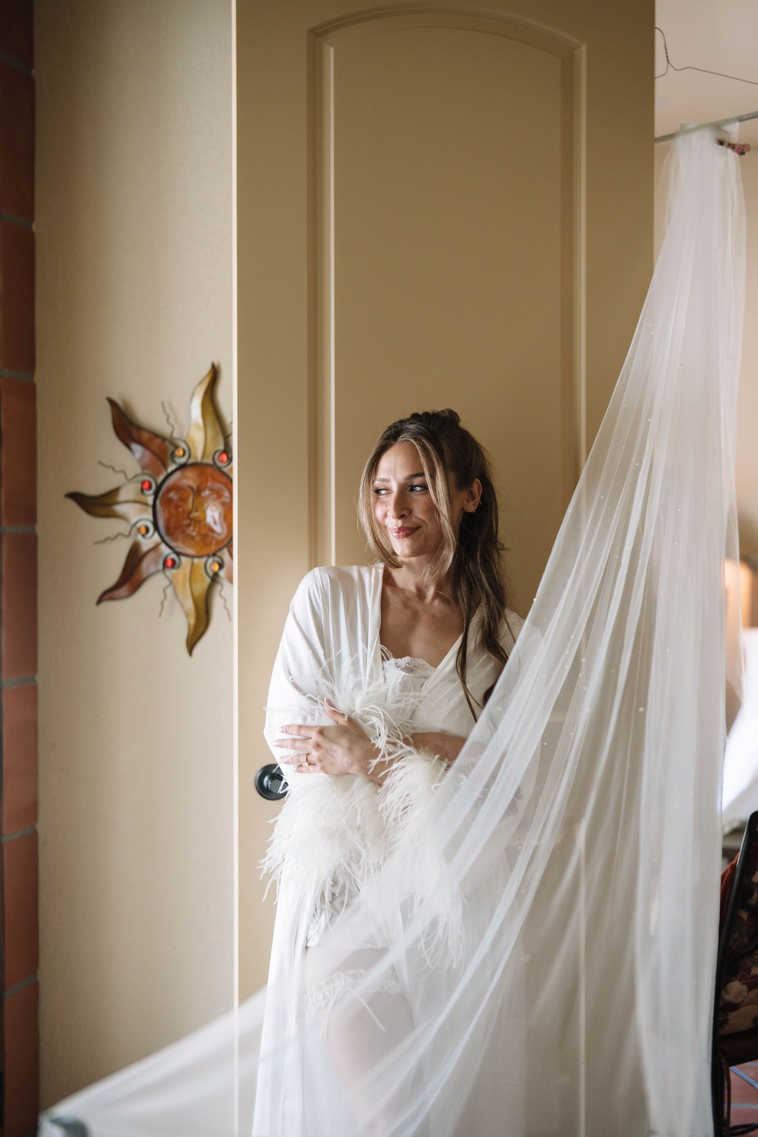 Bride in white robe holding feather bouquet stands by door with sun wall decor, smiling softly in warm light.