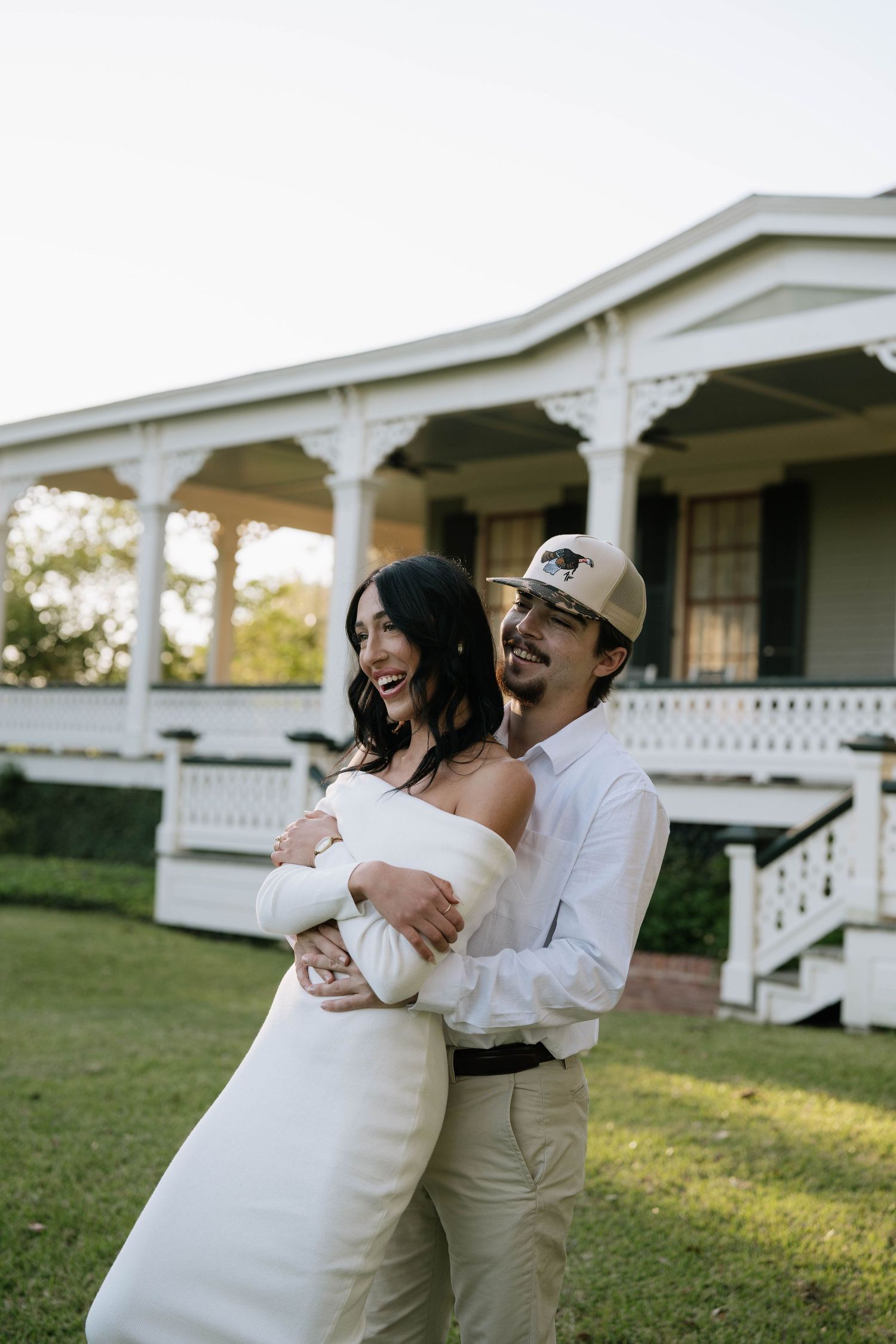 Couple embracing and laughing in front of a white Victorian porch, bride in off-shoulder gown, groom in cap and white shirt.
