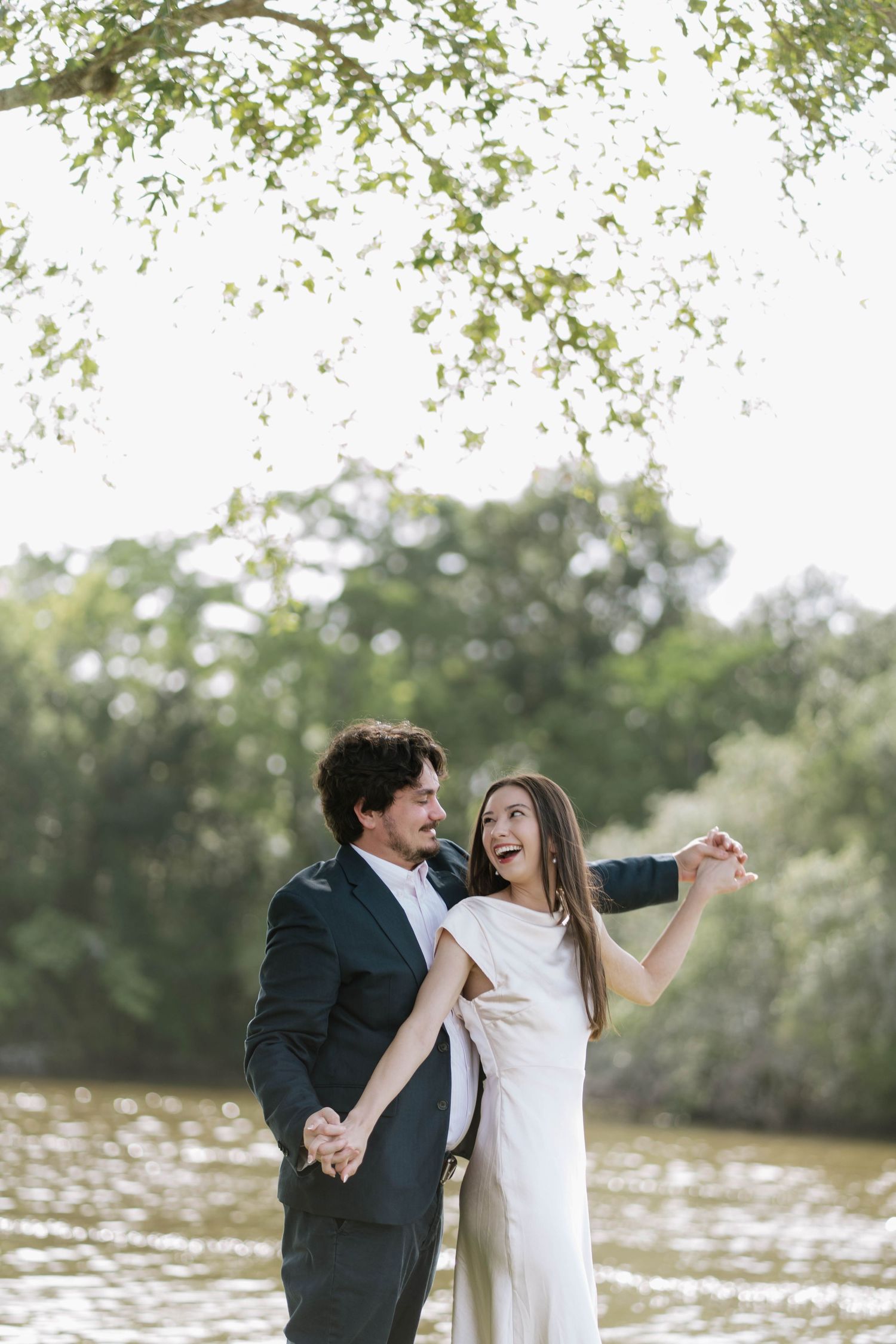 Joyful bride in white dress and groom in dark suit holding hands outdoors near a river surrounded by lush green trees.