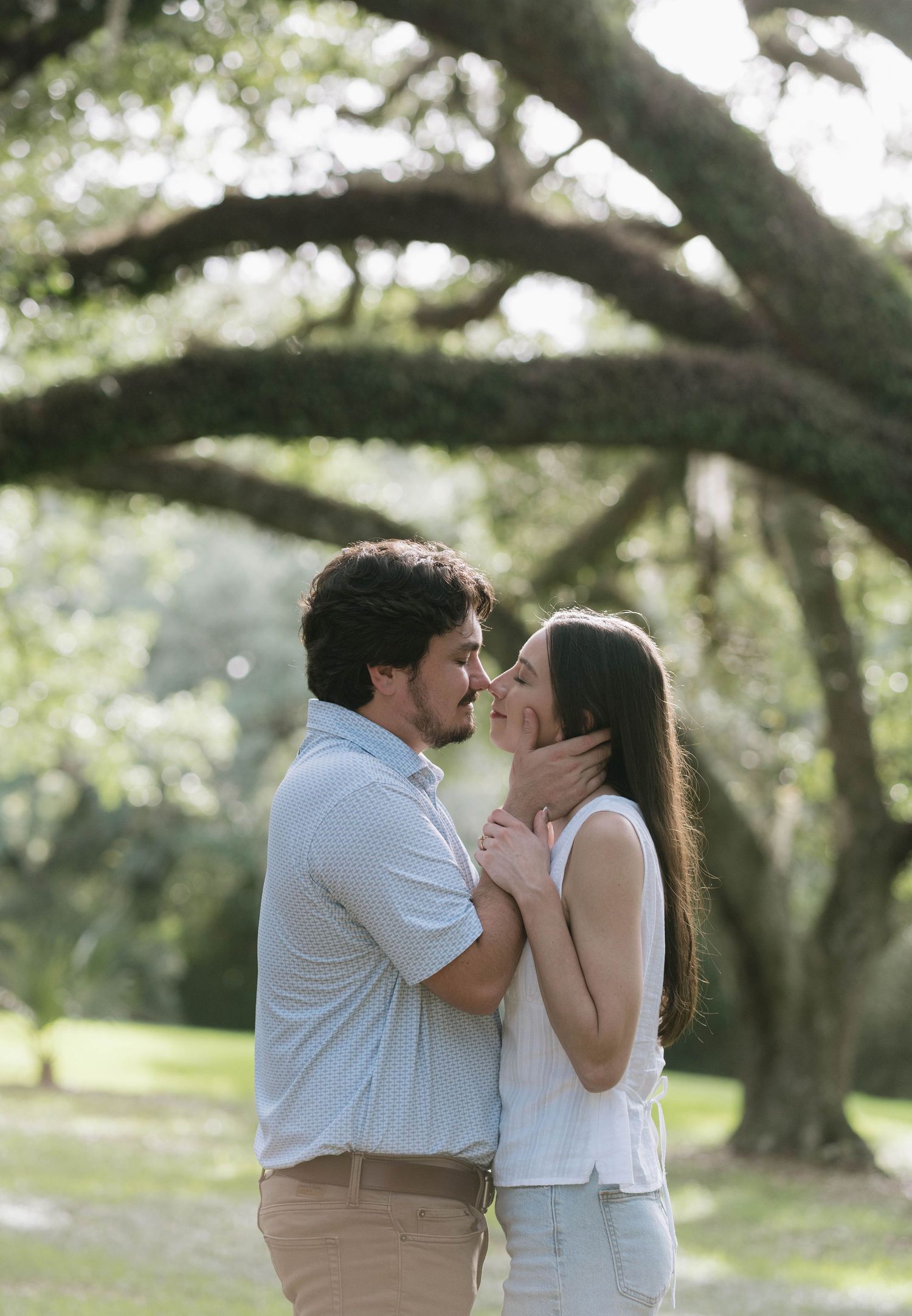 Couple sharing a romantic almost-kiss moment outdoors under large oak trees in a lush green park setting.